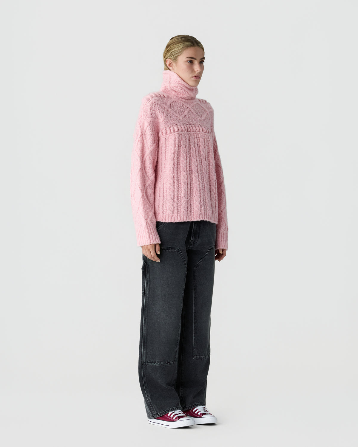 Person wearing a pink cable knit turtleneck sweater, dark wide leg jeans, and red and white sneakers against a light gray background