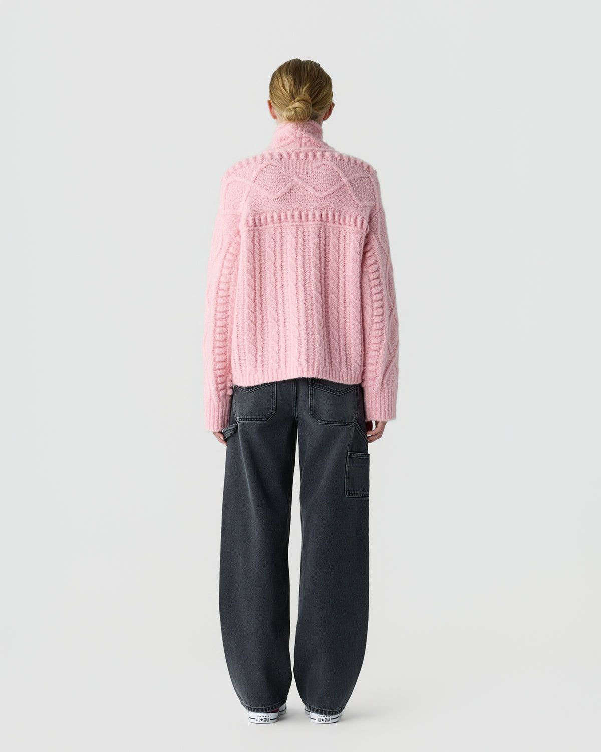 Back view of a person wearing a pink textured knit sweater with a high neck and black wide leg trousers against a light gray background