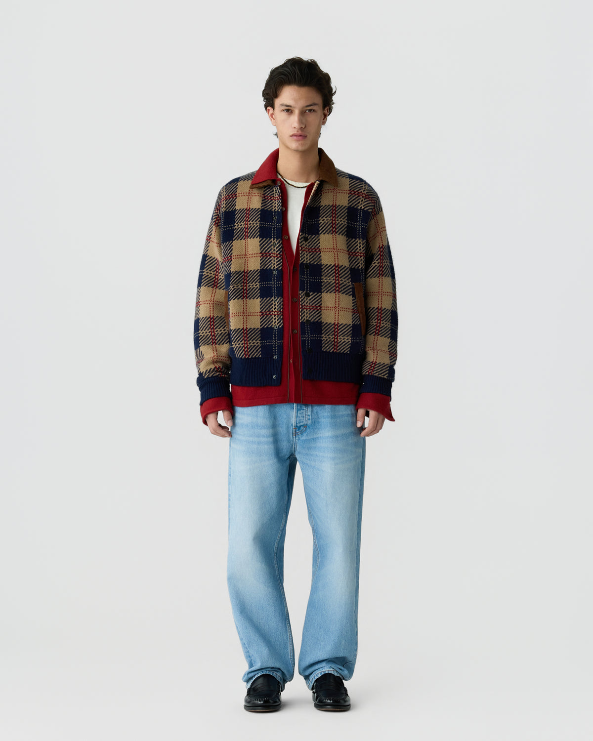 Young man wearing a beige, navy, and red plaid jacket over a white shirt paired with light blue jeans and black shoes, standing against a plain white background.