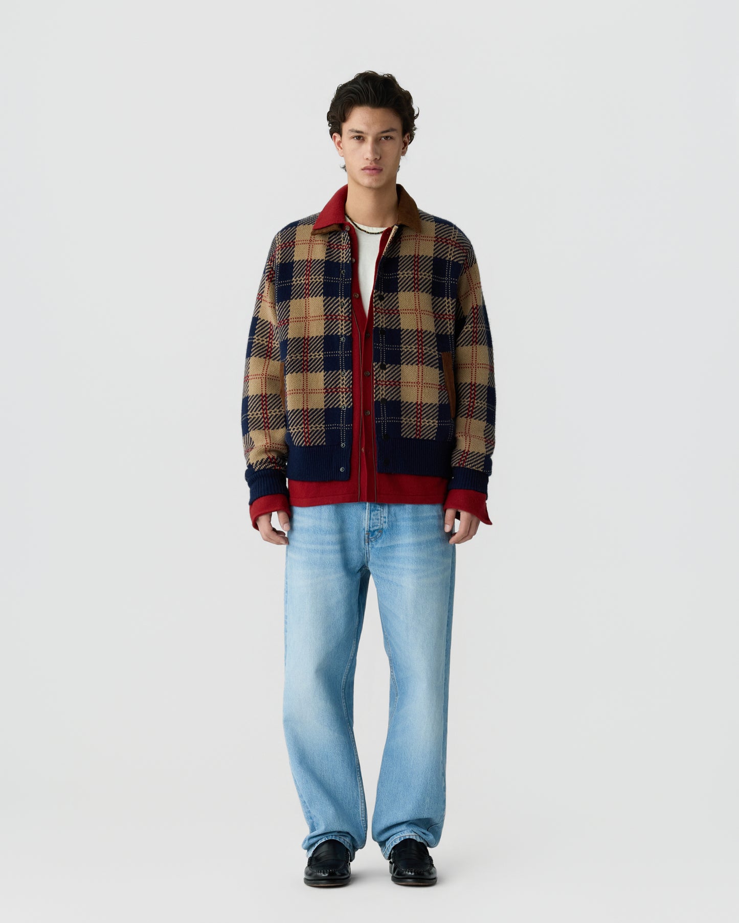 Young man wearing a beige, navy, and red plaid jacket over a white shirt paired with light blue jeans and black shoes, standing against a plain white background.