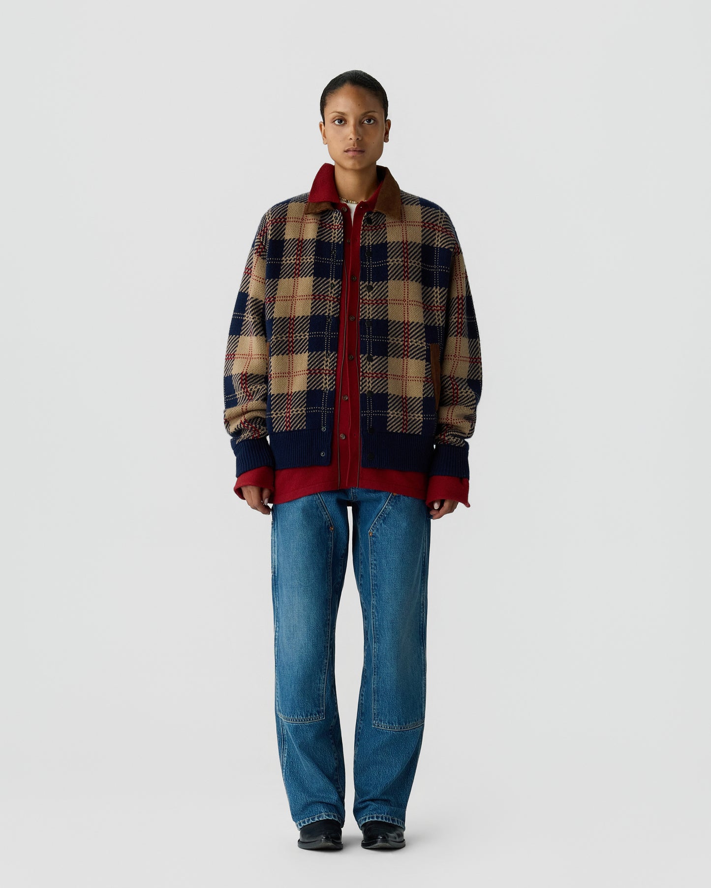 Person wearing a beige, navy, and red plaid jacket over a red zip-up hoodie, paired with blue jeans and black shoes, standing against a plain light gray background.