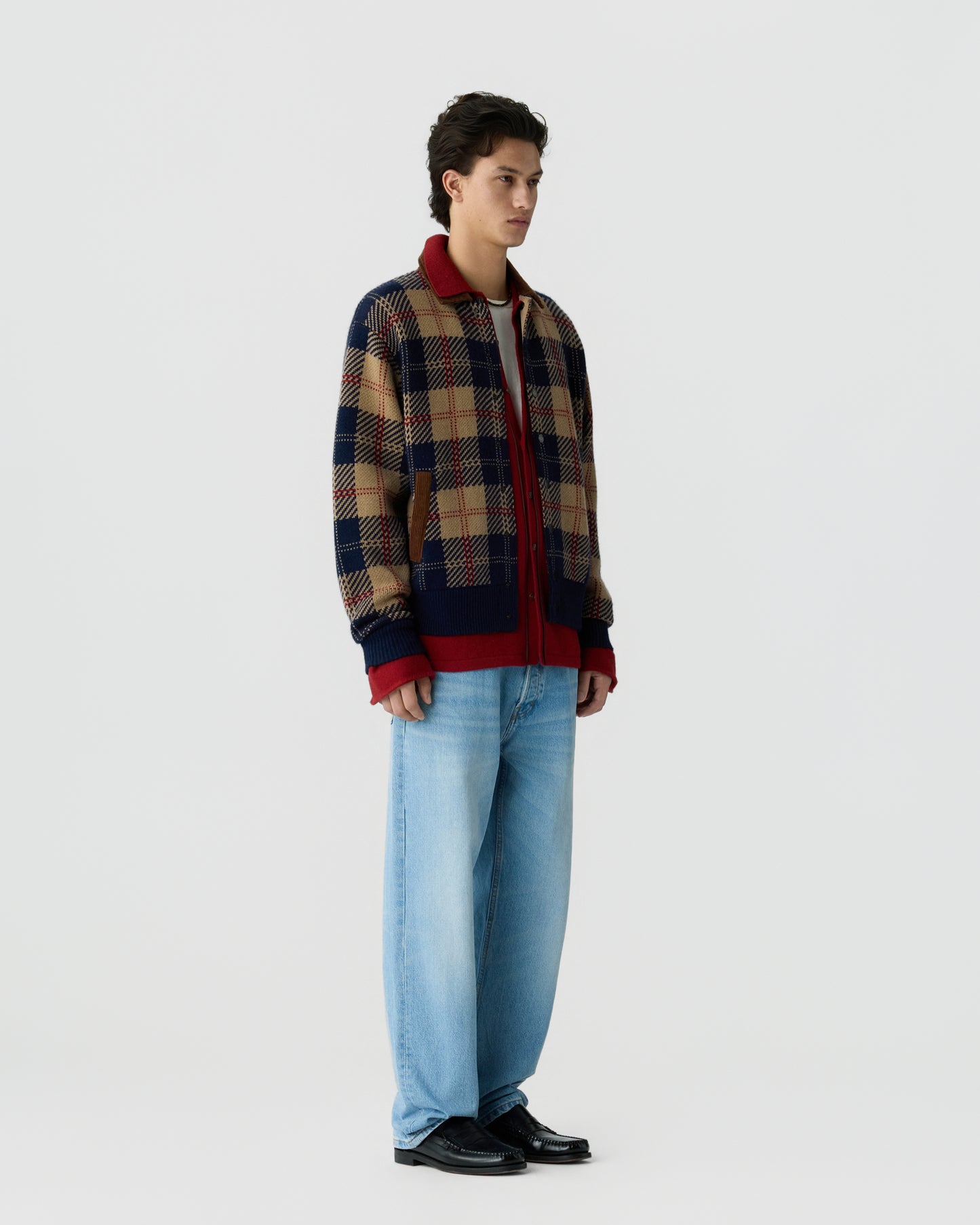 Young man wearing a navy and tan plaid jacket with red accents, light blue wide-leg jeans, and black shoes, standing against a plain light gray background.