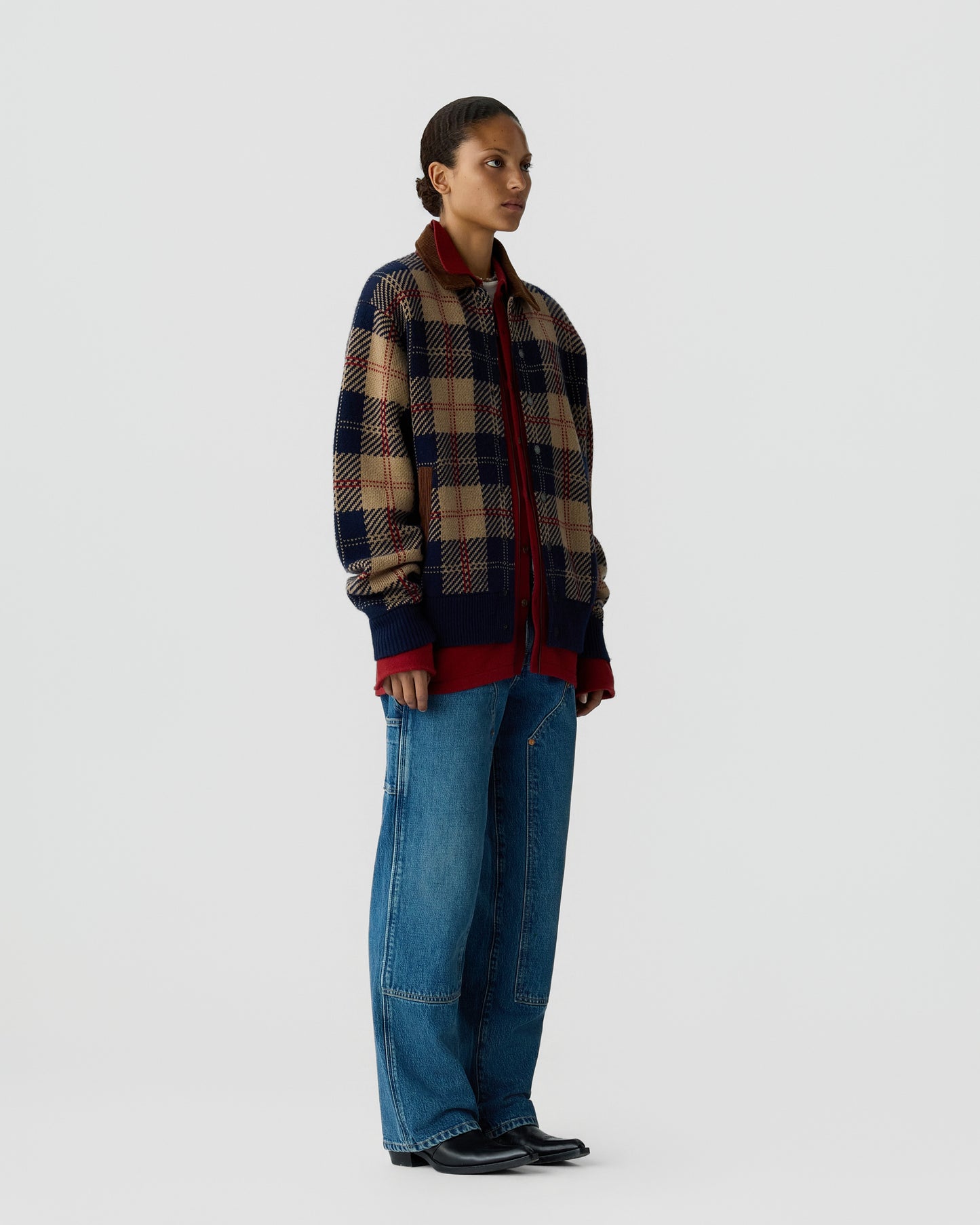 Person wearing a blue, red, and beige plaid jacket over a red shirt, paired with wide-leg blue jeans and black shoes, standing against a plain light background.