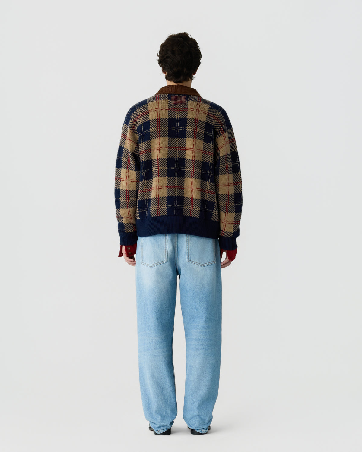 Man standing facing away wearing a beige and navy plaid sweater with red accents paired with light blue jeans against a plain background.