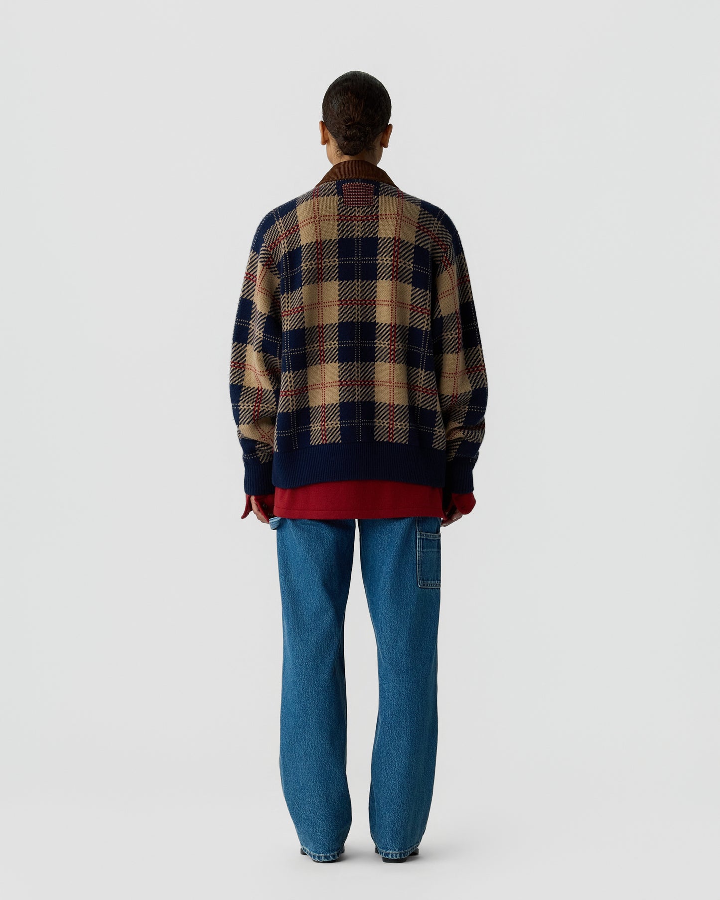 Person wearing a navy and beige plaid sweater with a dark brown collar, red shirt underneath, blue jeans, and red gloves, facing away against a plain background.