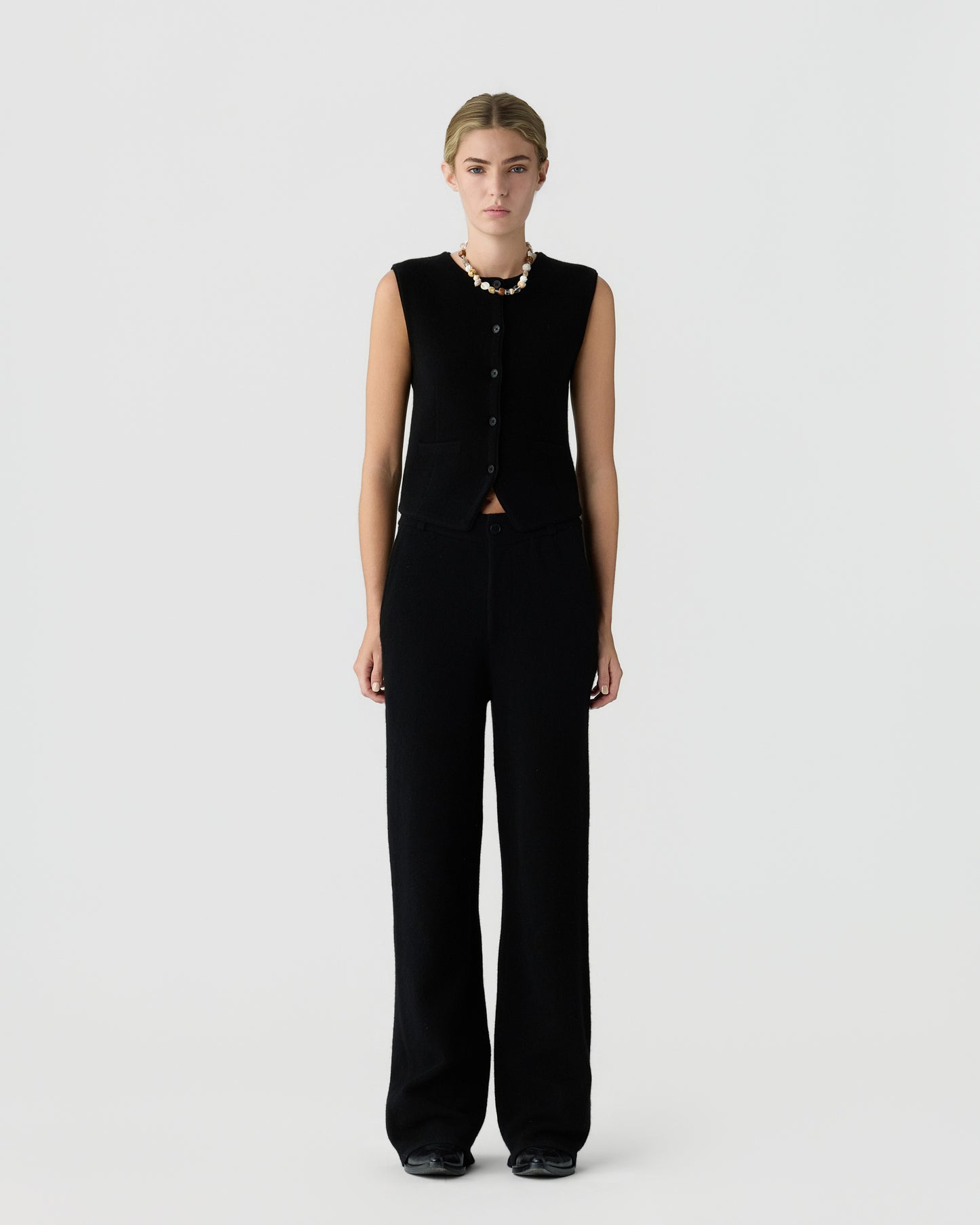 Young woman wearing a sleeveless black button-up vest and matching wide-leg black pants, accessorized with a chunky white and beige necklace, standing against a plain white background.