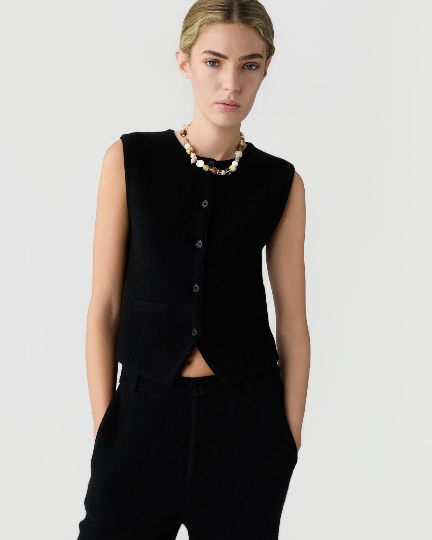 Young woman wearing a sleeveless black button-up vest and matching black pants against a plain light background, accessorized with a chunky beaded necklace