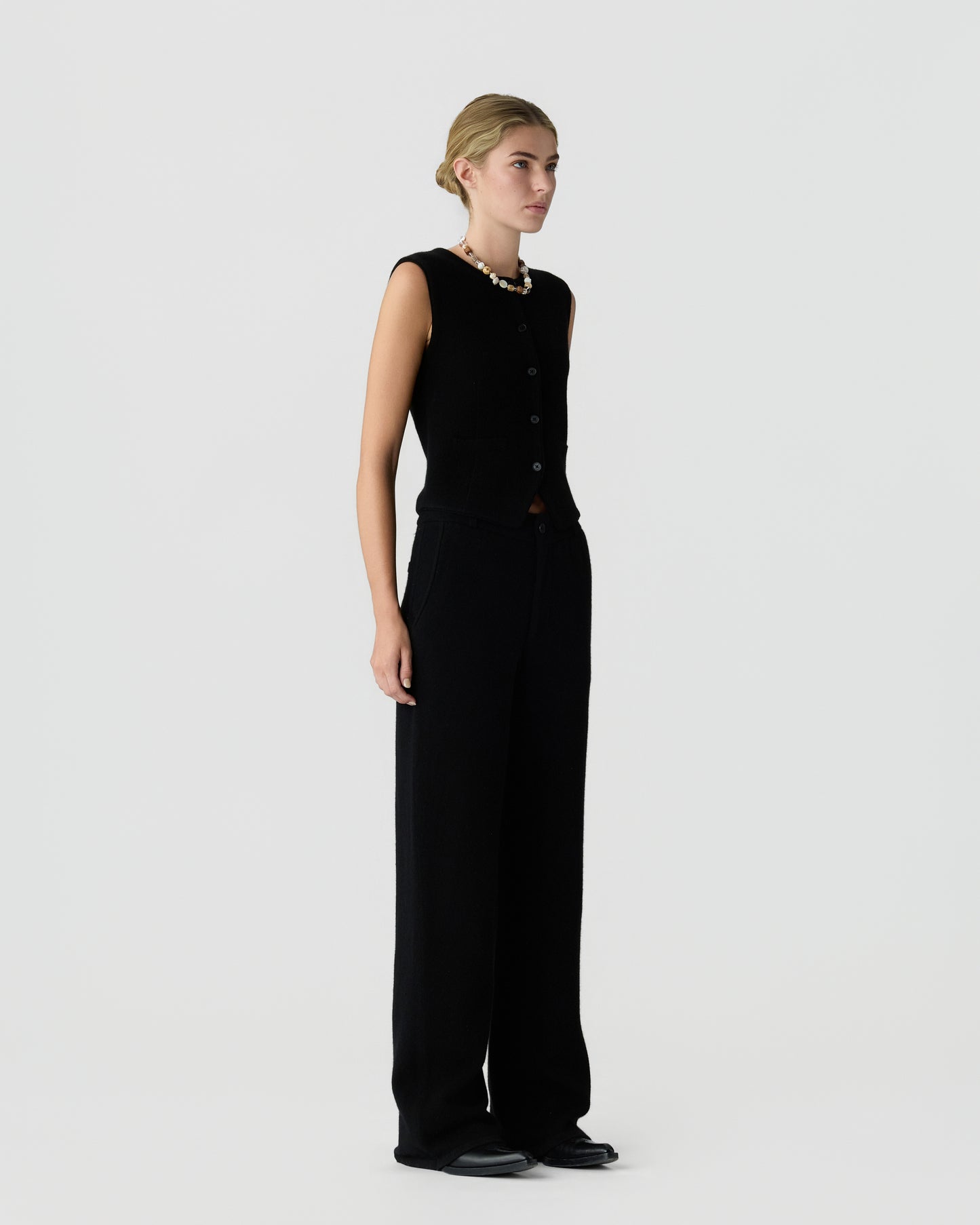 Side view of a woman wearing a sleeveless black buttoned vest and wide-leg black pants, standing against a plain light background with a necklace around her neck.
