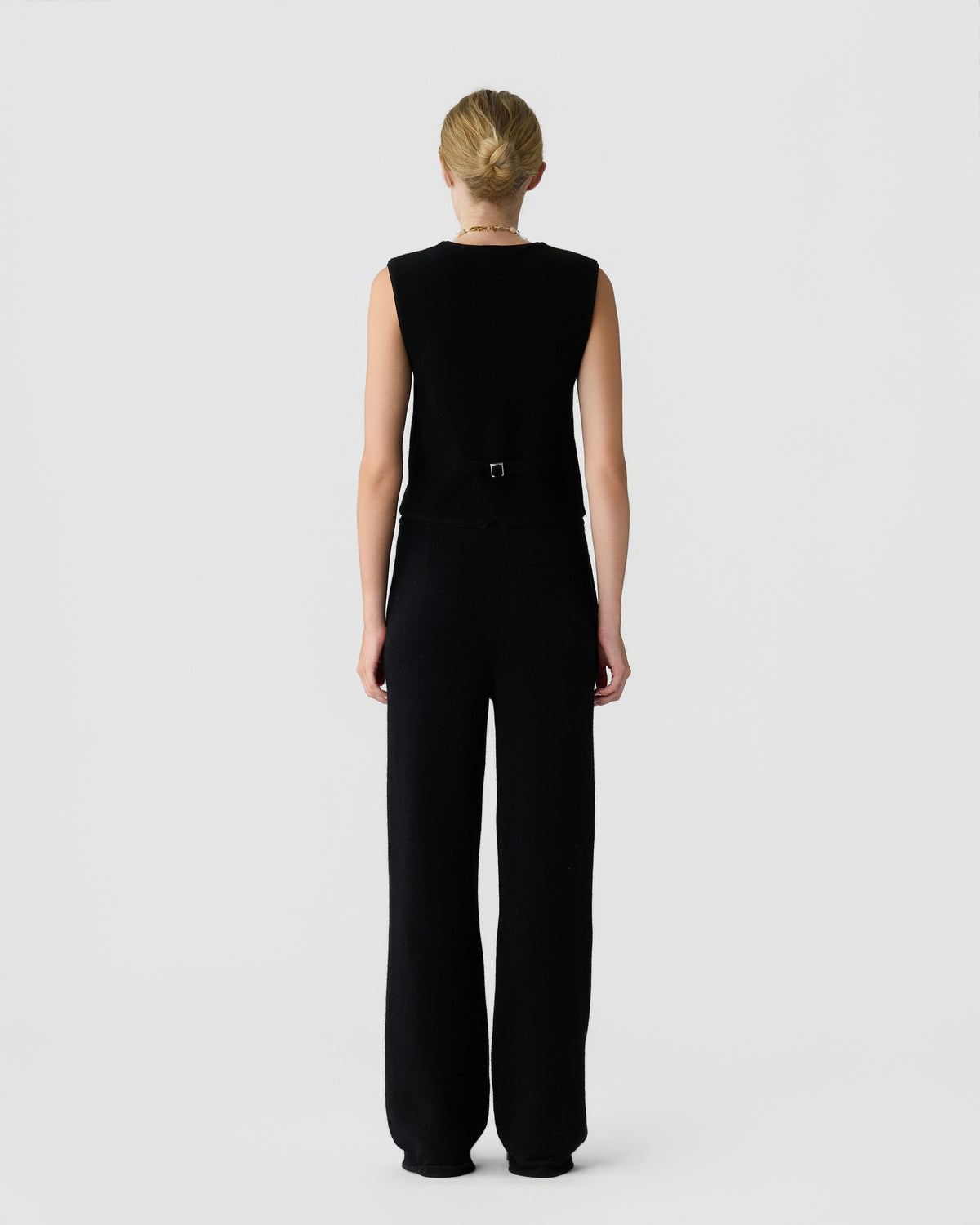 Woman with blonde hair tied in a bun wearing a sleeveless black top and wide-leg black pants standing facing away against a plain white background.