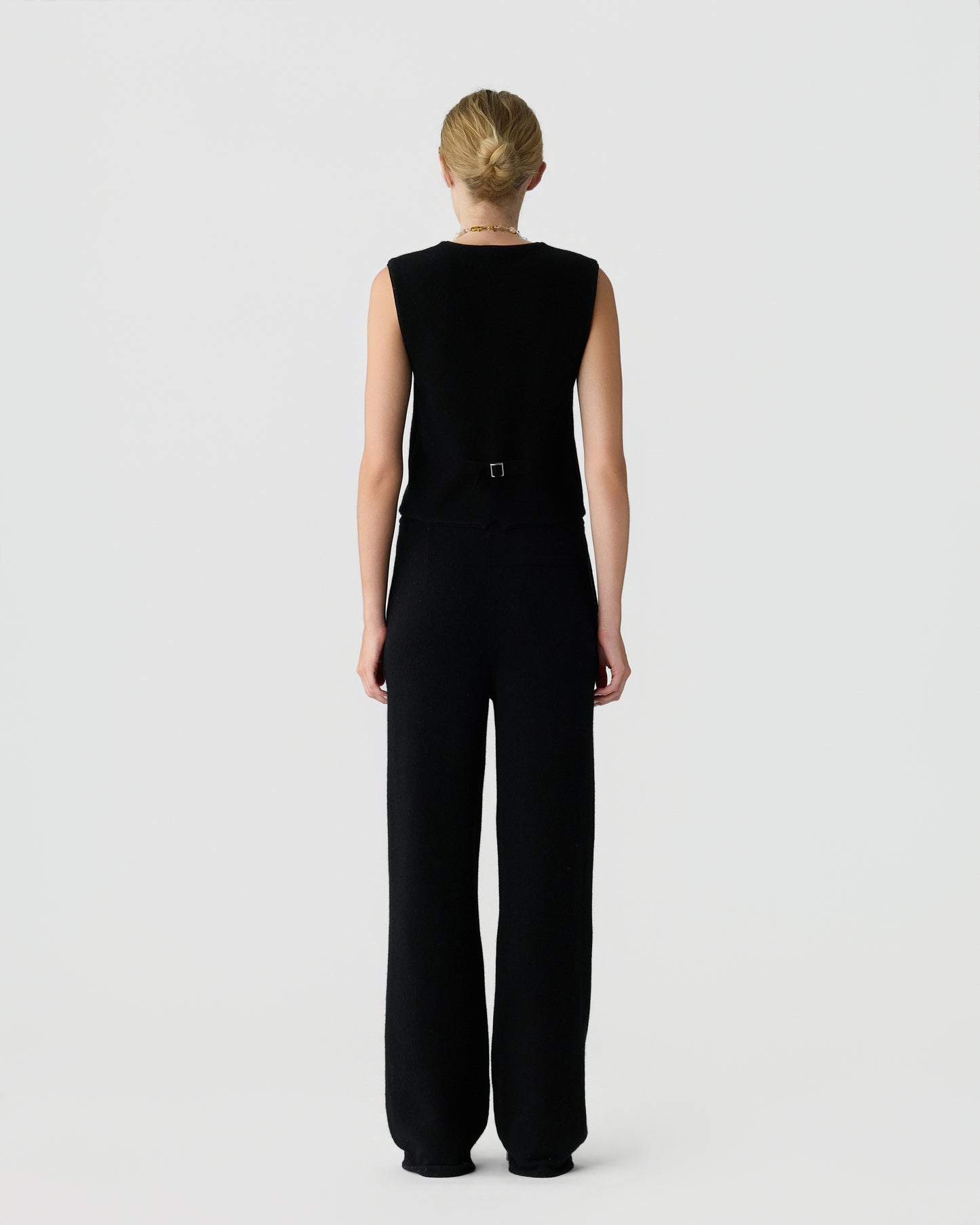 Woman with blonde hair tied in a bun wearing a sleeveless black top and wide-leg black pants standing facing away against a plain white background.