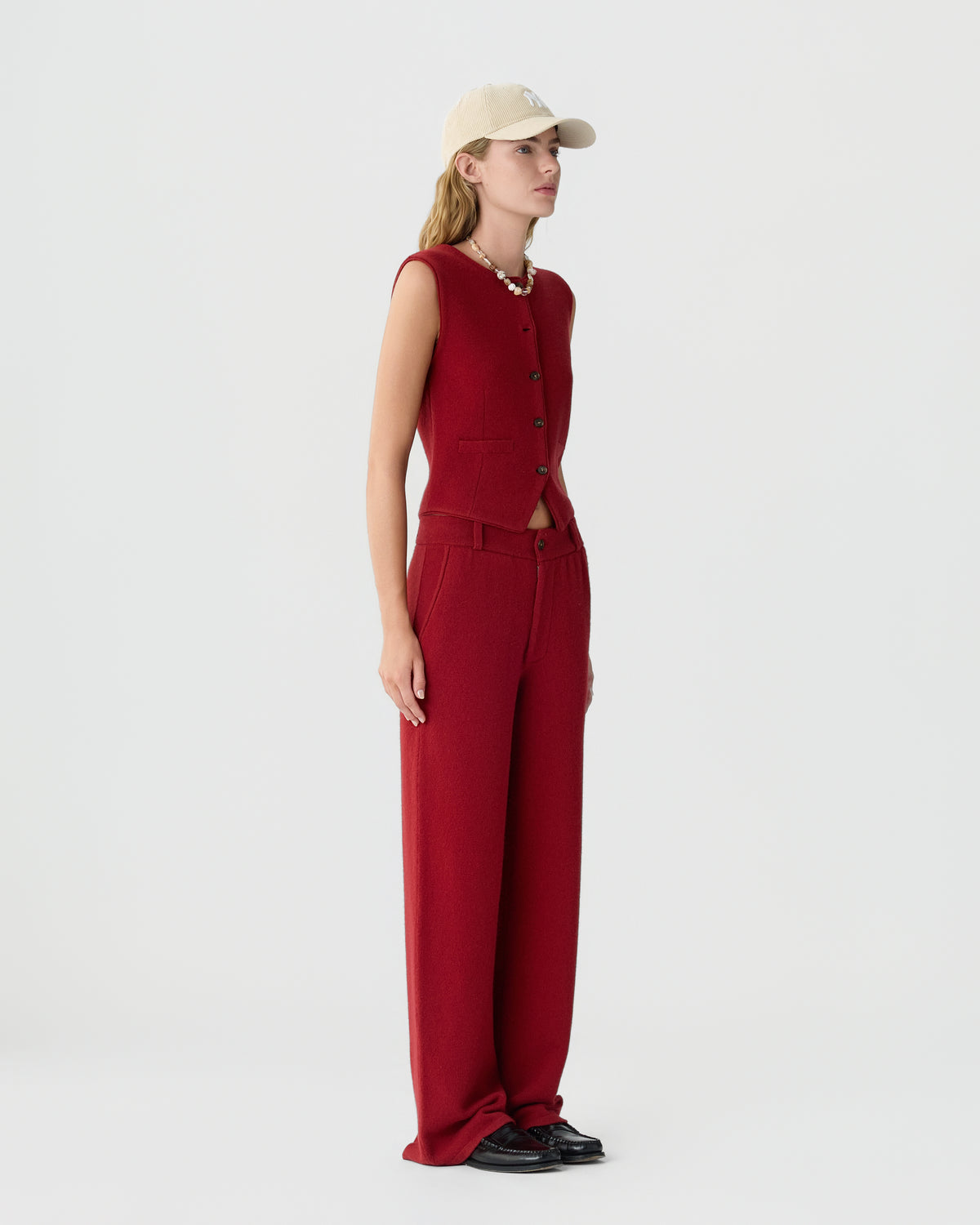 Woman wearing a sleeveless red buttoned vest and matching wide-leg pants, paired with a beige baseball cap, black shoes, and a chunky necklace against a plain white background.