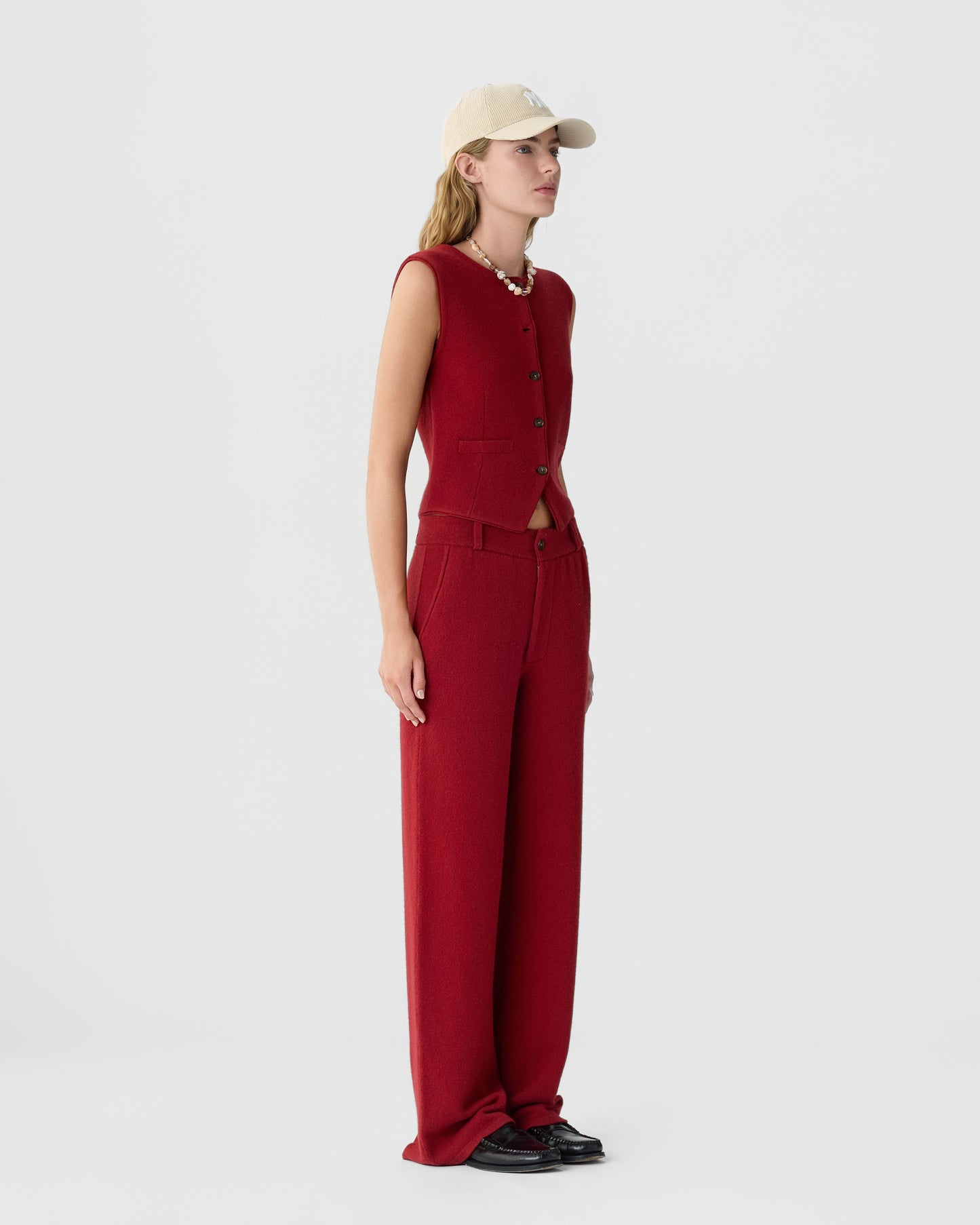 Woman wearing a sleeveless red buttoned vest and matching wide-leg pants, paired with a beige baseball cap, black shoes, and a chunky necklace against a plain white background.