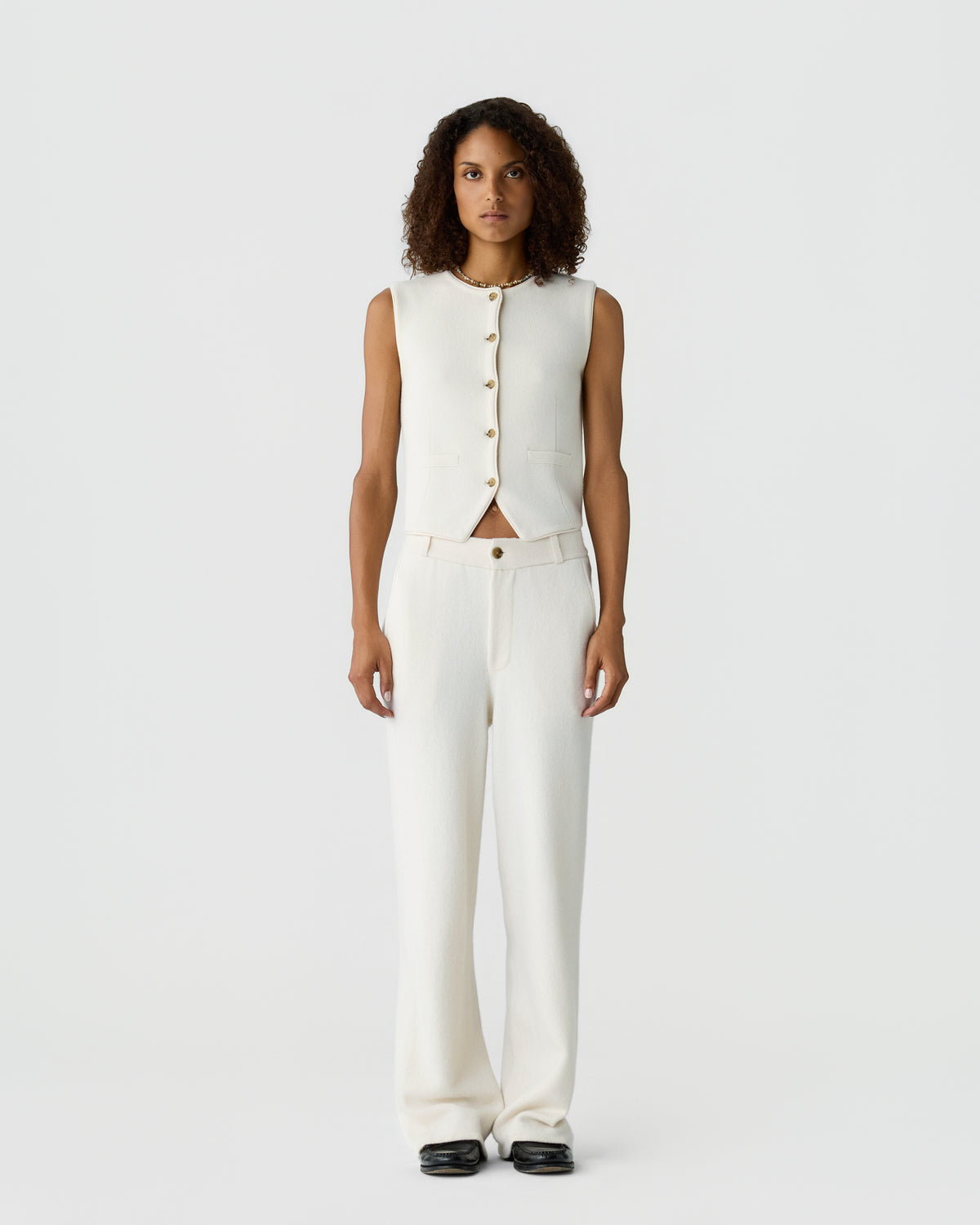 A woman with curly hair stands against a plain white background wearing a sleeveless, buttoned cream vest and matching cream wide-leg pants with black shoes.
