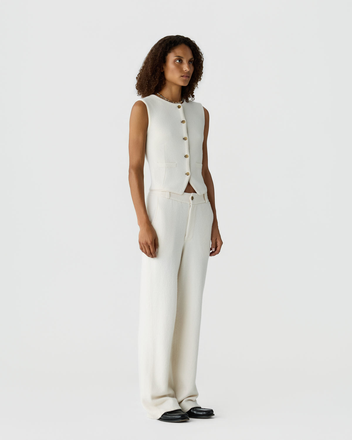 Woman with curly hair wearing a sleeveless white buttoned vest and matching wide-leg trousers standing against a plain light background with black shoes.