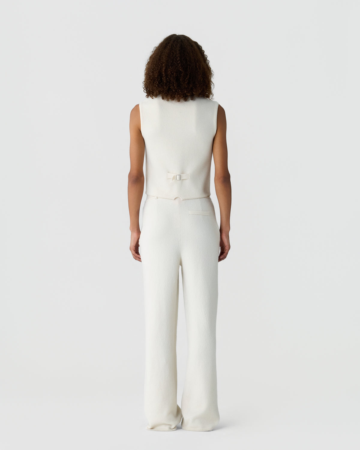 Woman with curly hair wearing a sleeveless white top with a back buckle and matching white wide-leg pants, standing against a plain light gray background.