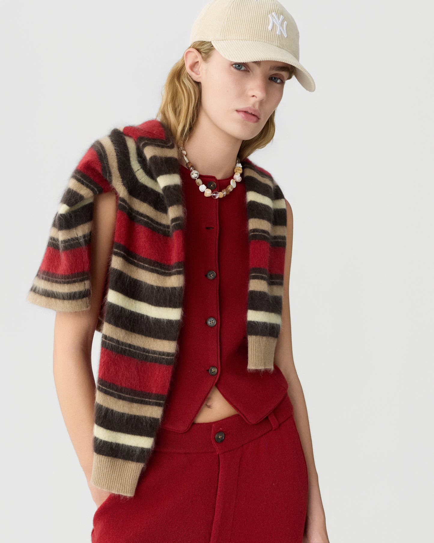 Young woman wearing a beige baseball cap, a red sleeveless button-up top, matching red pants, and a striped red, brown, black, and beige cashmere sweater draped over her shoulders with a chunky necklace.
