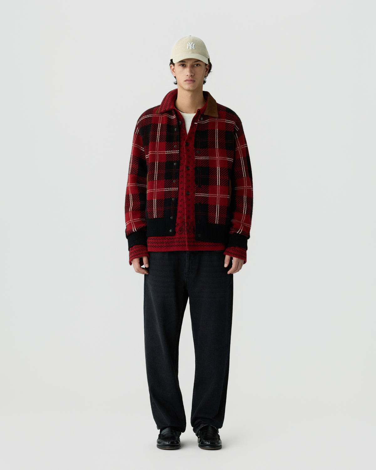 Young man wearing a red and black plaid jacket over a red patterned shirt, black pants, black shoes, and a beige baseball cap standing against a plain white background.