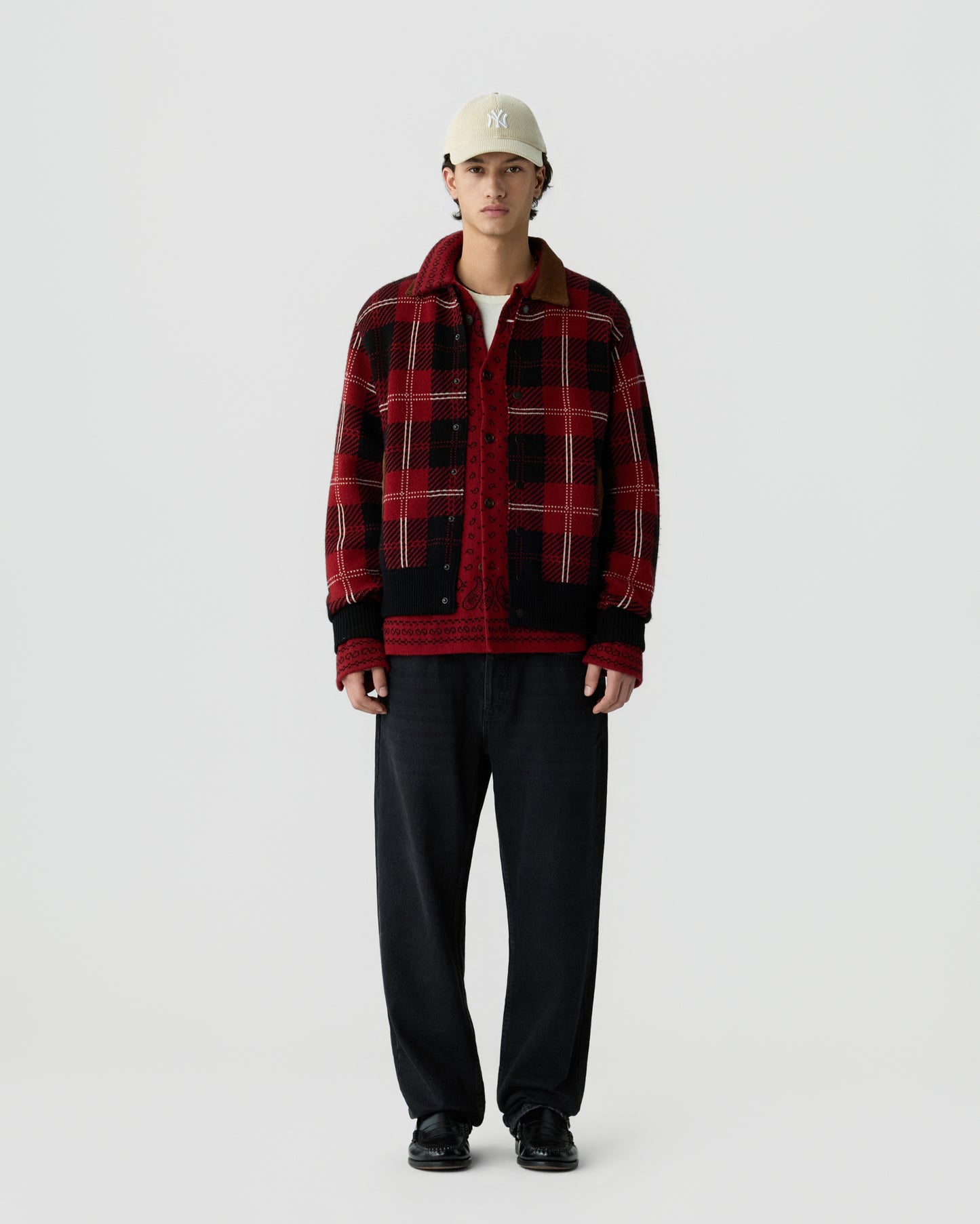 Young man wearing a red and black plaid jacket over a red patterned shirt, black pants, black shoes, and a beige baseball cap standing against a plain white background.