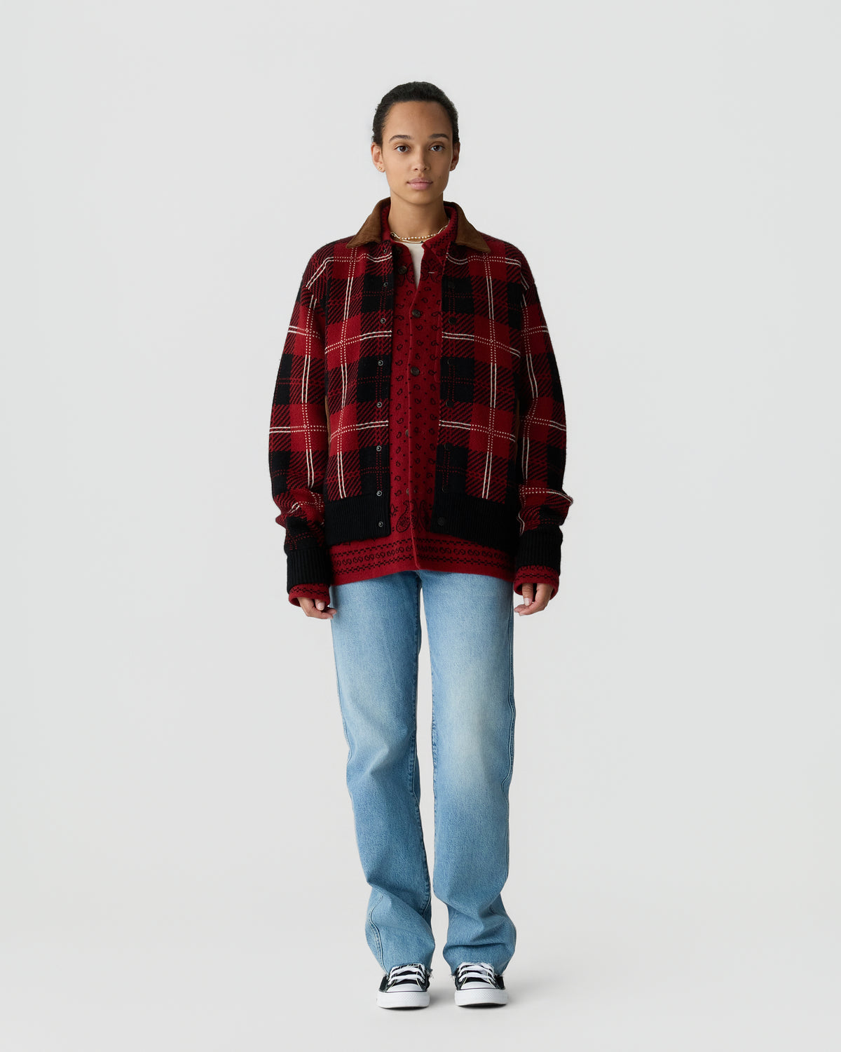 A person stands against a plain white background wearing a red and black plaid jacket over a red knitted sweater, light blue jeans, and black and white sneakers.
