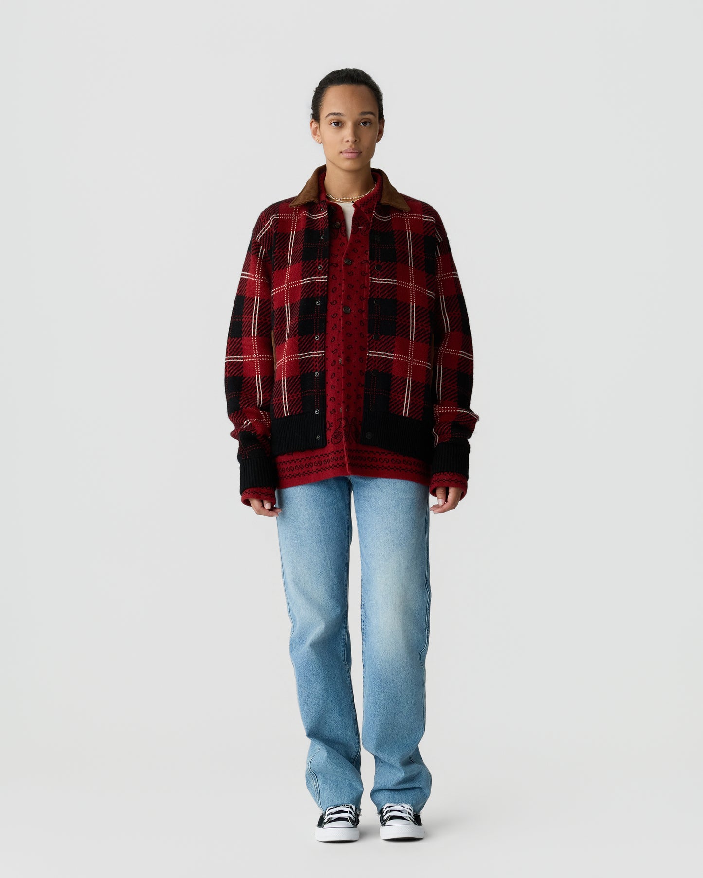A person stands against a plain white background wearing a red and black plaid jacket over a red knitted sweater, light blue jeans, and black and white sneakers.