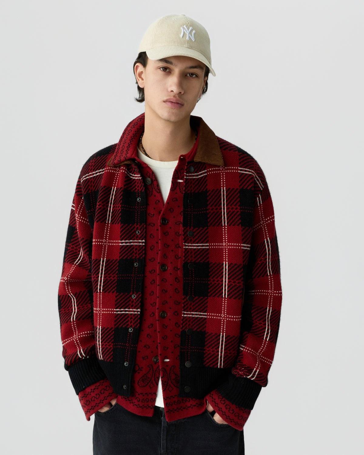 Young man wearing a beige baseball cap, white T-shirt, red patterned button-up shirt, and a red and black checkered jacket with black cuffs and a brown collar against a plain gray background.