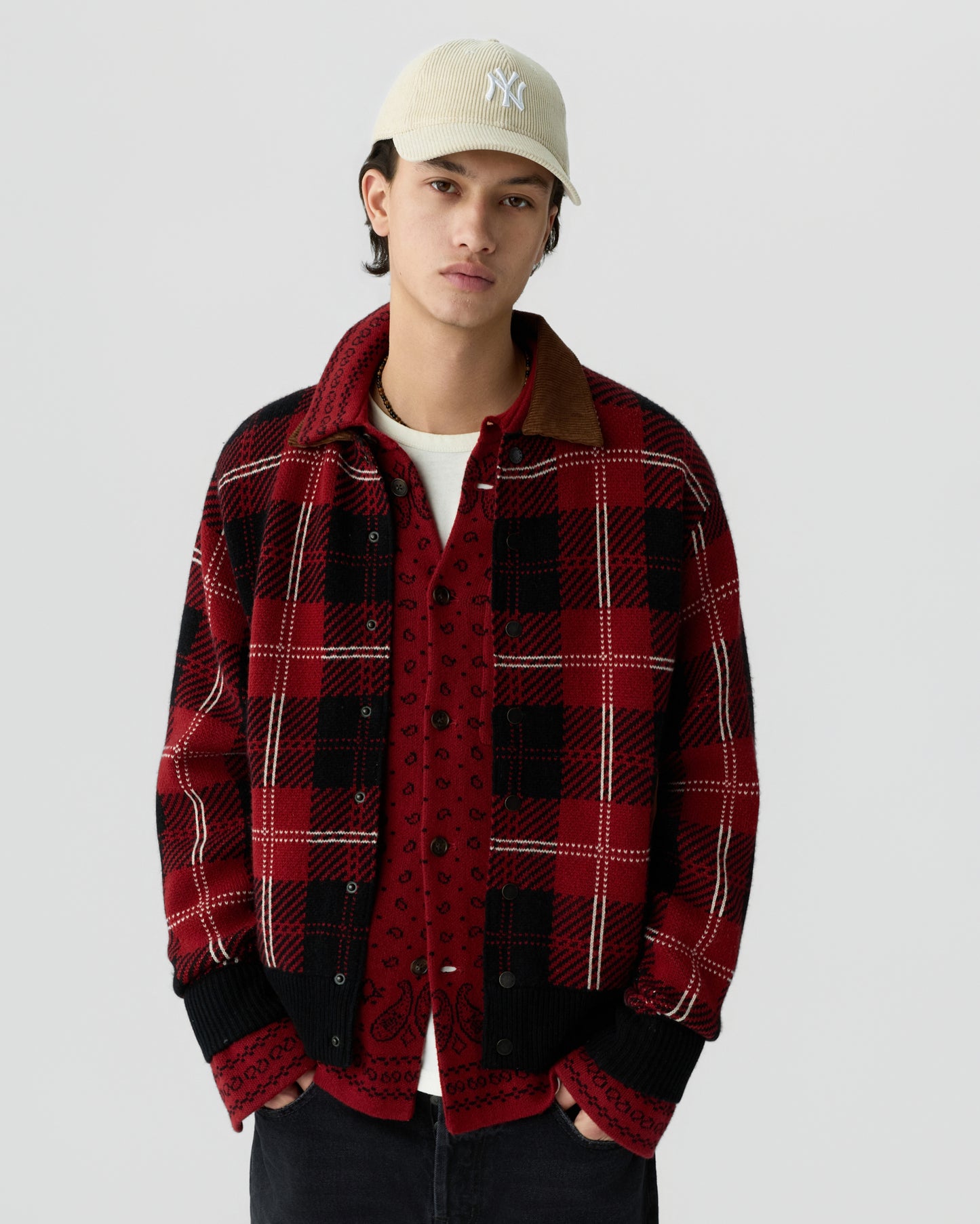 Young man wearing a beige baseball cap, white T-shirt, red patterned button-up shirt, and a red and black checkered jacket with black cuffs and a brown collar against a plain gray background.