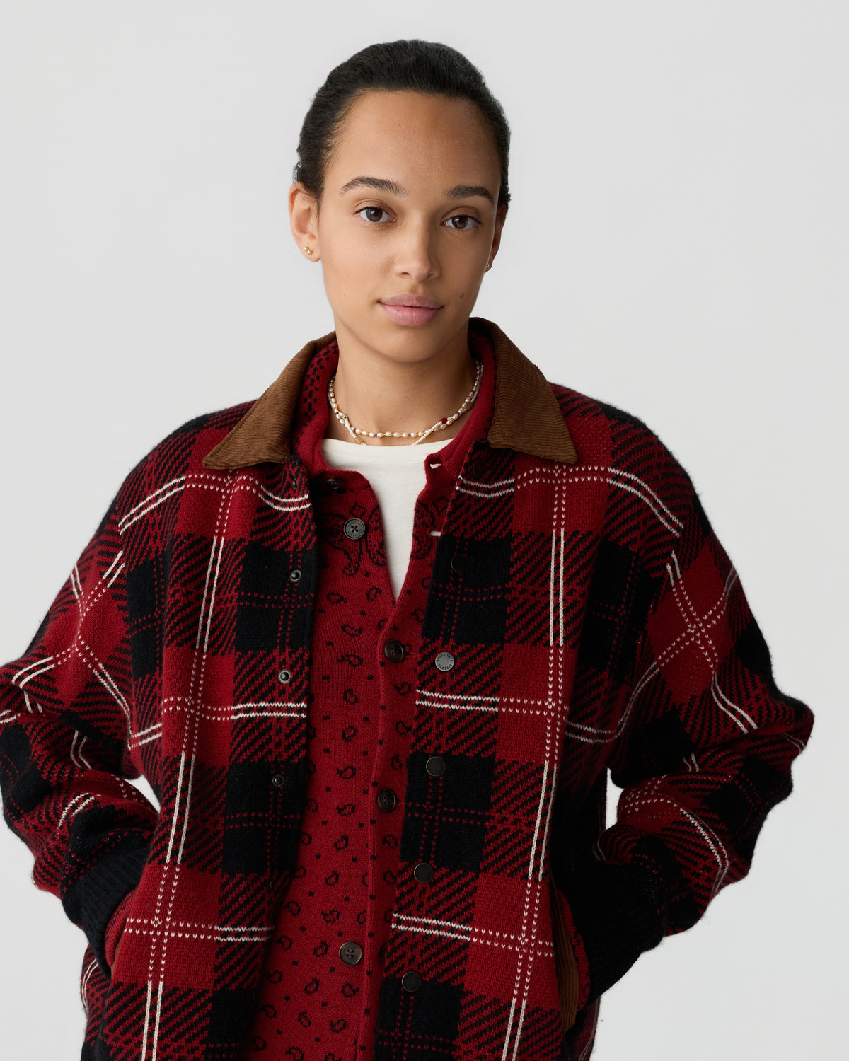 A young woman with dark hair pulled back wears a red and black plaid jacket with a brown collar over a red patterned shirt, posing against a plain light background.