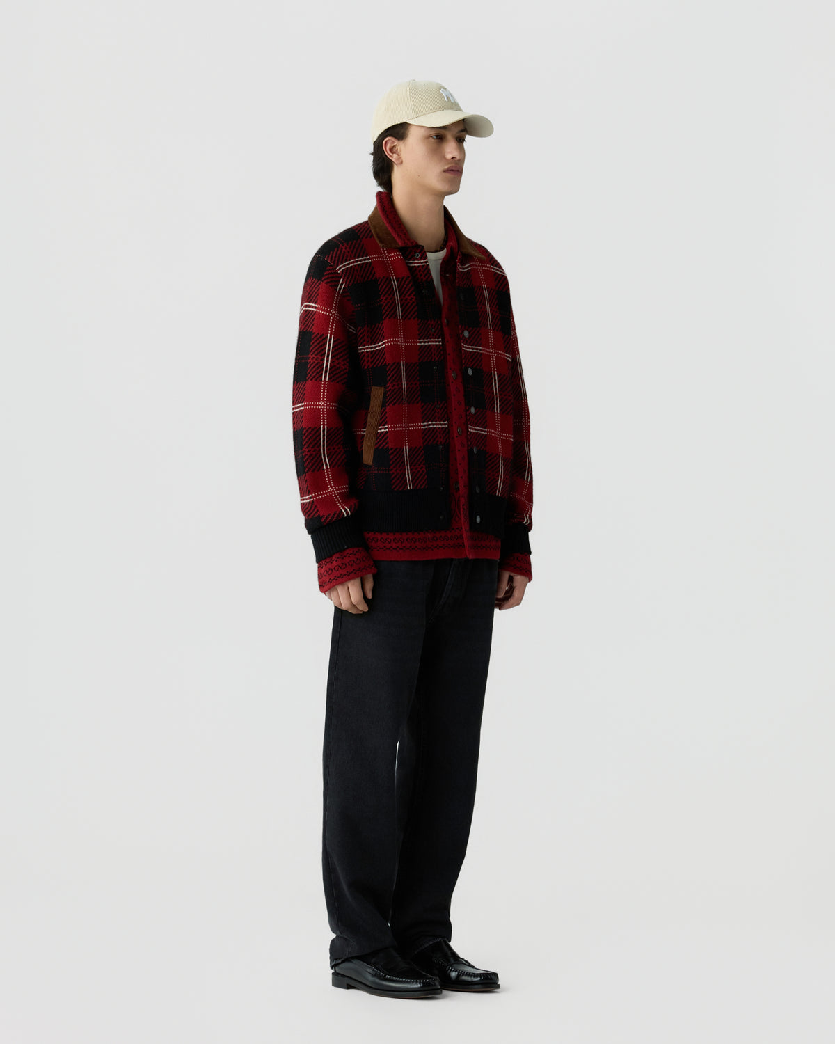 A young man wearing a red and black plaid jacket, black pants, black shoes, and a cream-colored baseball cap standing against a plain white background.