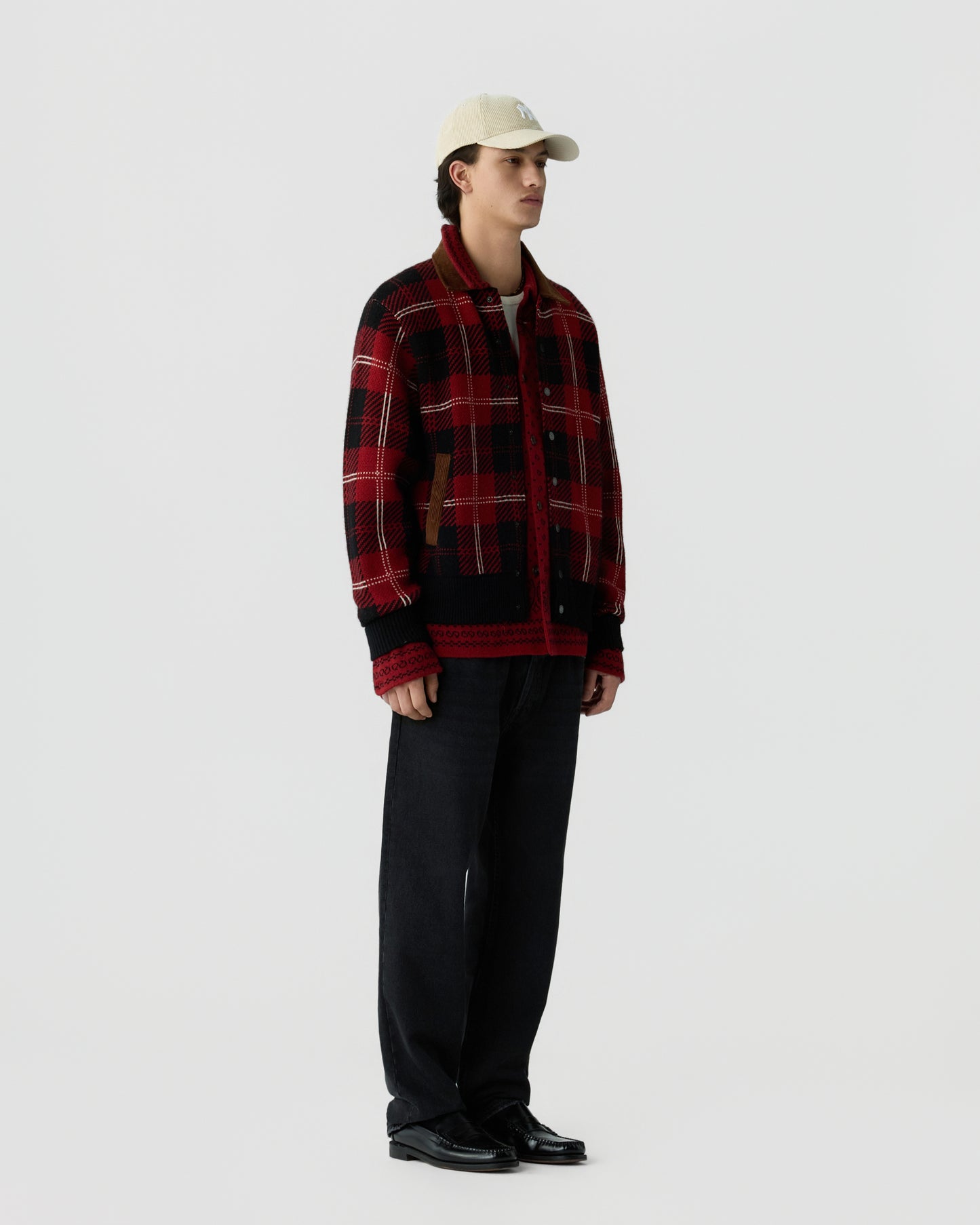 A young man wearing a red and black plaid jacket, black pants, black shoes, and a cream-colored baseball cap standing against a plain white background.