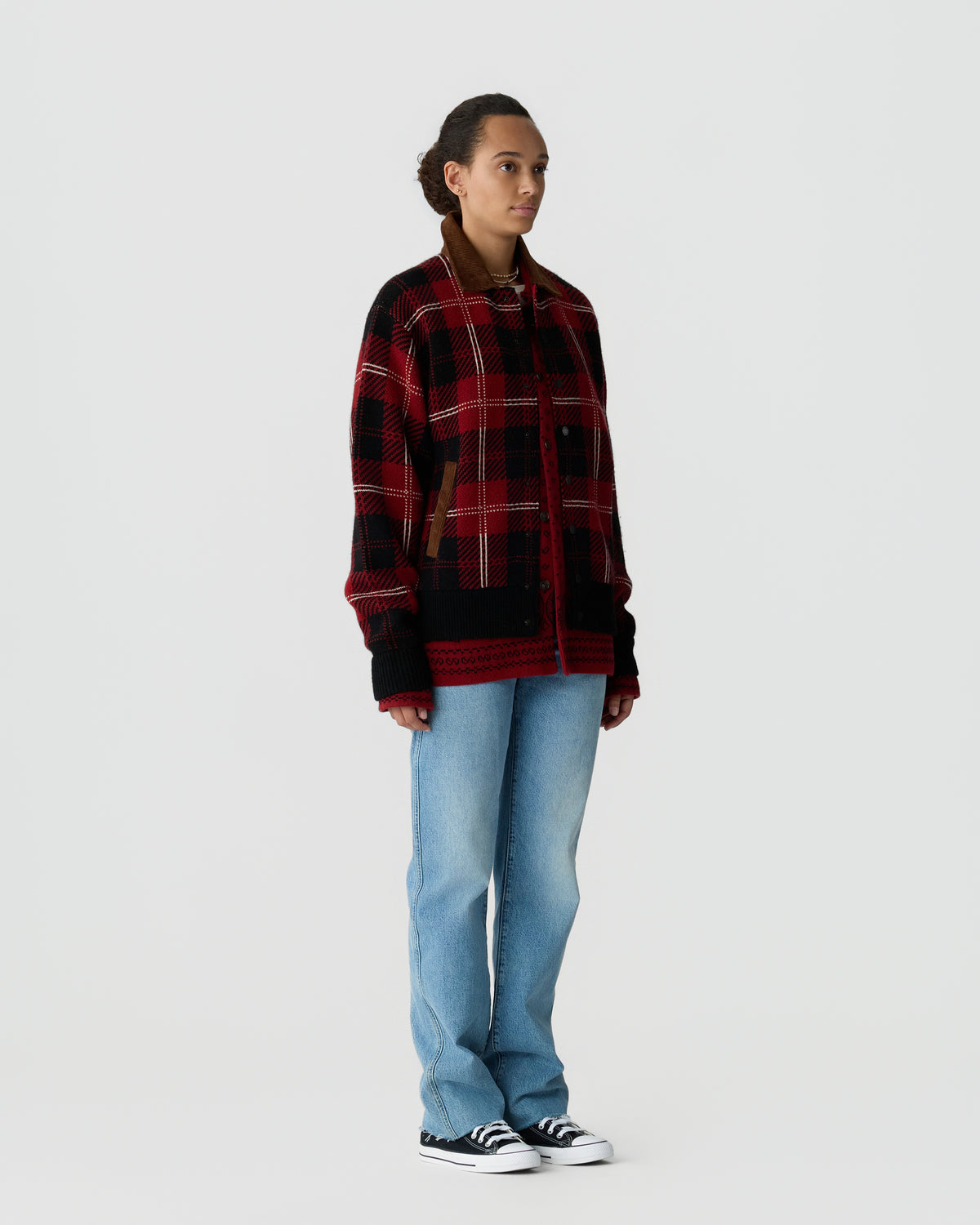 Woman standing wearing a red and black plaid jacket with brown collar details, light blue jeans, and black and white sneakers against a plain light background.