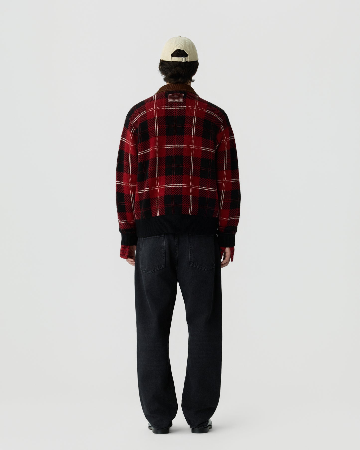 Person wearing a red and black plaid jacket, dark jeans, and a light-colored baseball cap, standing facing away against a plain white background.
