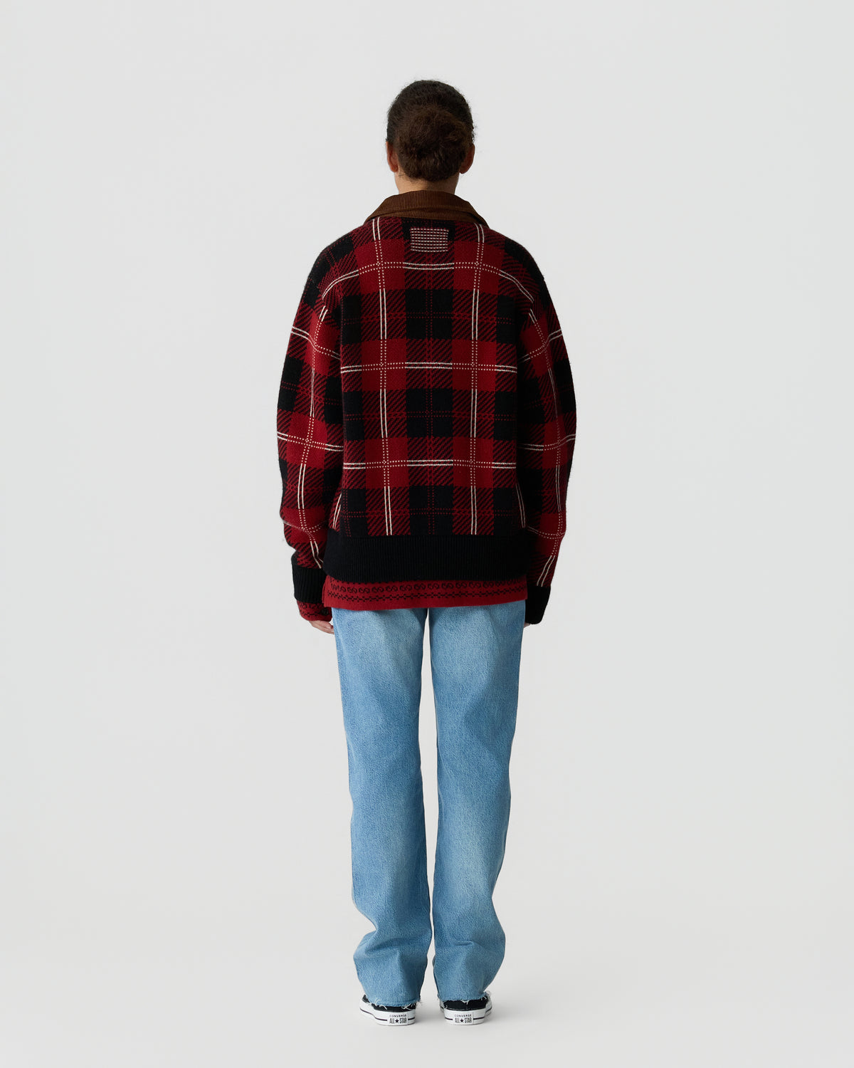 Person wearing a red and black plaid oversized sweater with black cuffs, light blue jeans, and black and white sneakers, standing facing away against a plain background.