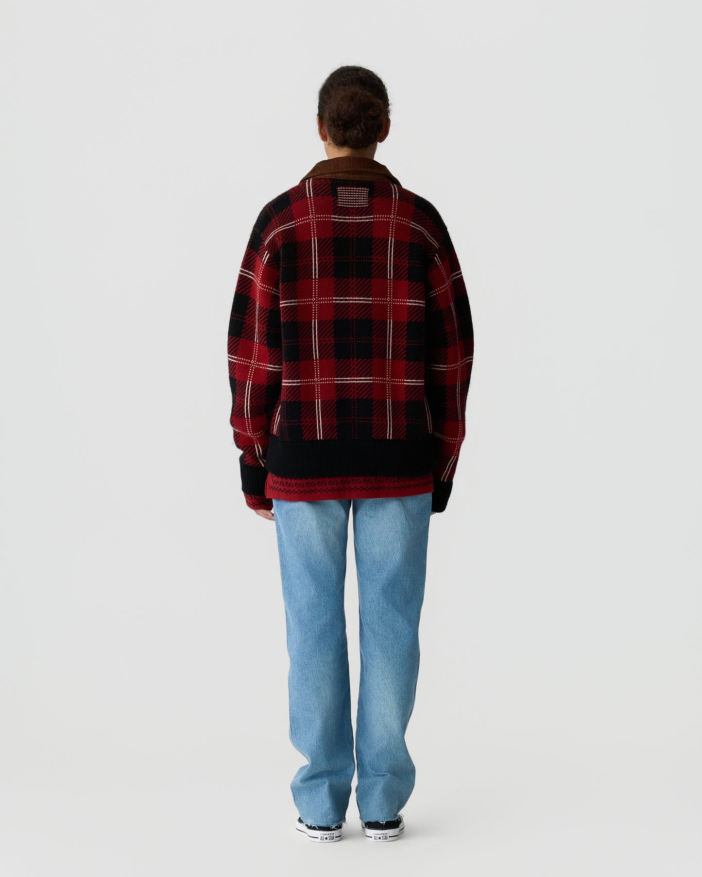 Person wearing a red and black plaid oversized sweater with black cuffs, light blue jeans, and black and white sneakers, standing facing away against a plain background.