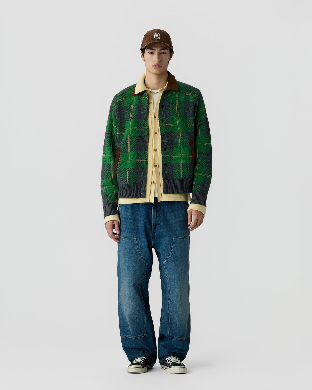 Young man wearing a green and black plaid jacket over a beige shirt, blue relaxed-fit jeans, black and white sneakers, and a brown baseball cap standing against a plain white background.