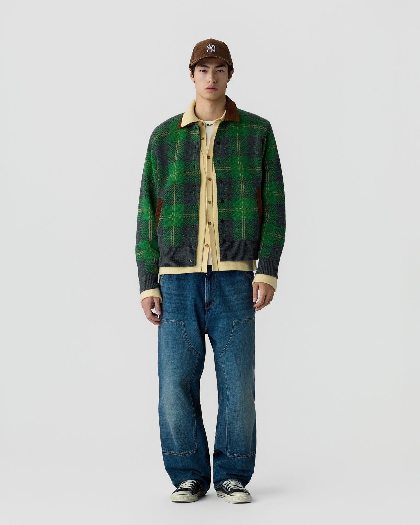 Young man wearing a green and black plaid jacket over a beige shirt, blue relaxed-fit jeans, black and white sneakers, and a brown baseball cap standing against a plain white background.