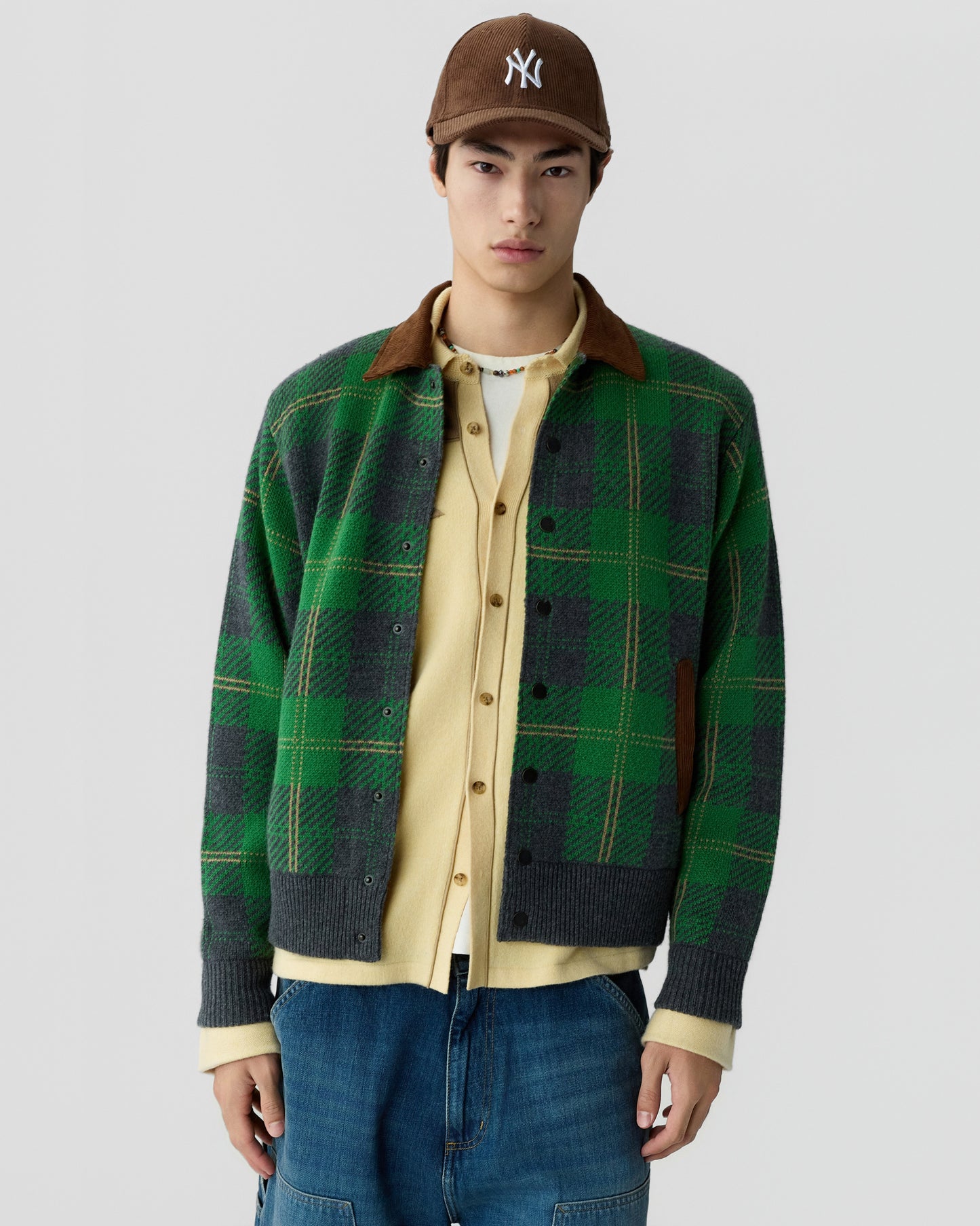 Young man wearing a green and grey plaid jacket over a beige button-up shirt and white t-shirt, paired with blue jeans and a brown baseball cap.