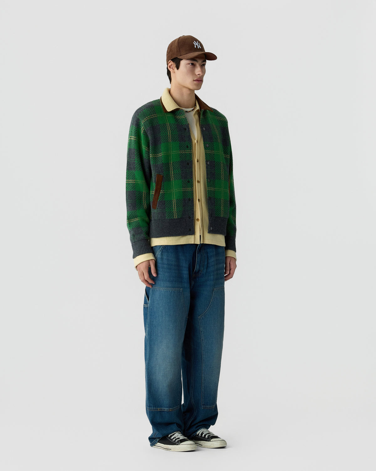 Young man wearing a brown baseball cap, layered clothing including a green and gray plaid jacket over a beige shirt, loose blue jeans, and black sneakers standing against a plain background.