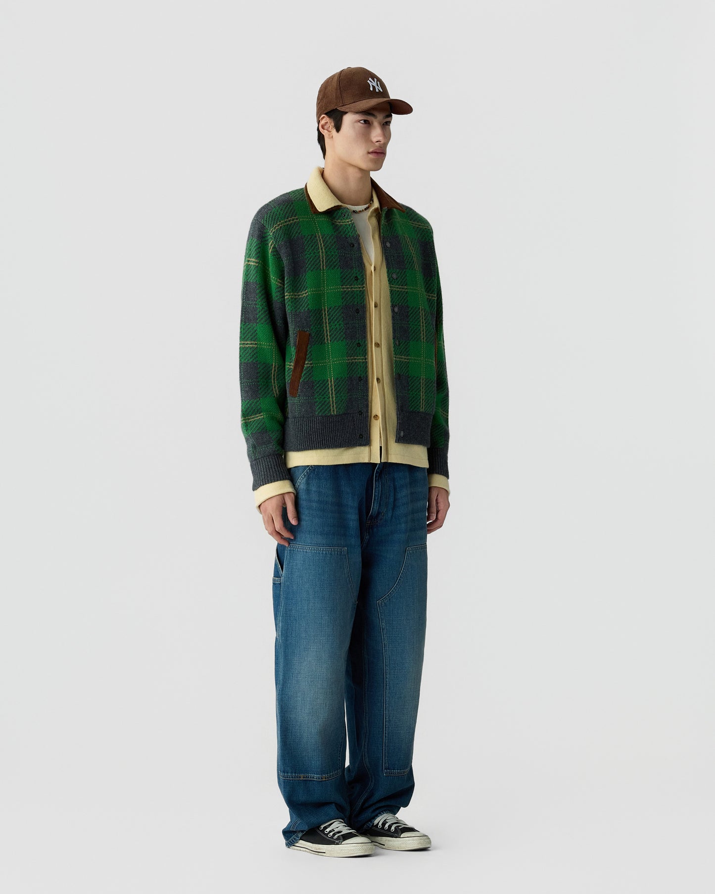 Young man wearing a brown baseball cap, layered clothing including a green and gray plaid jacket over a beige shirt, loose blue jeans, and black sneakers standing against a plain background.
