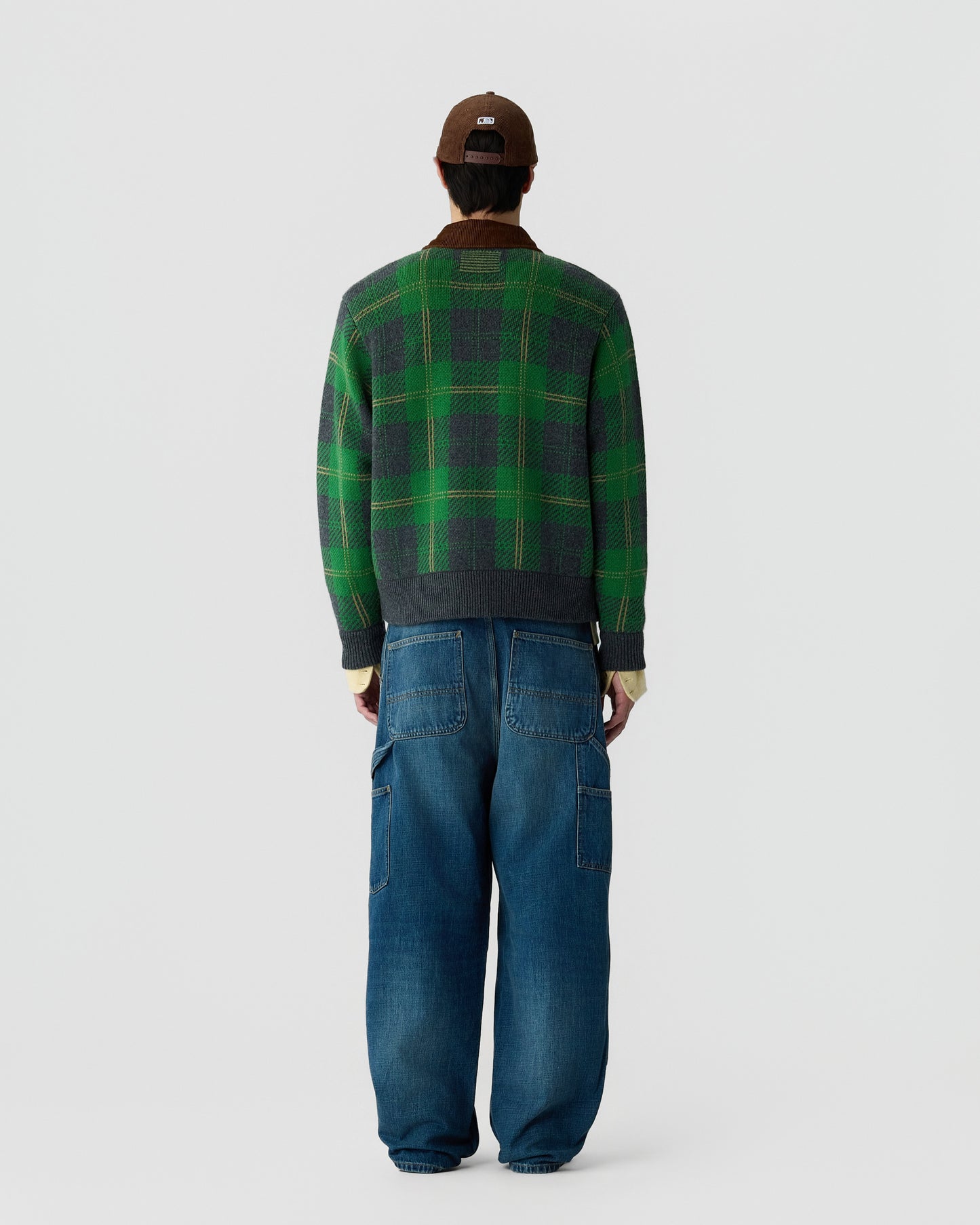 Man wearing a green and gray plaid sweater, loose-fitting blue denim jeans with pockets, a brown baseball cap, standing facing away against a plain background.