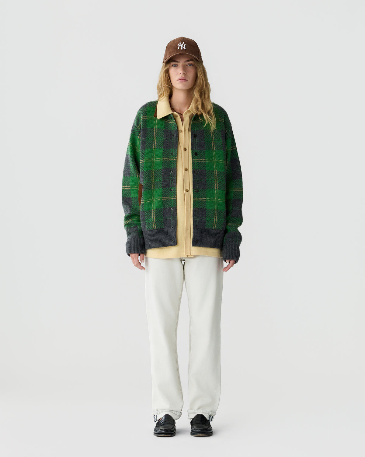 Young woman wearing a brown baseball cap, green and gray plaid cardigan over a yellow shirt, white pants, and black shoes, standing against a plain white background.