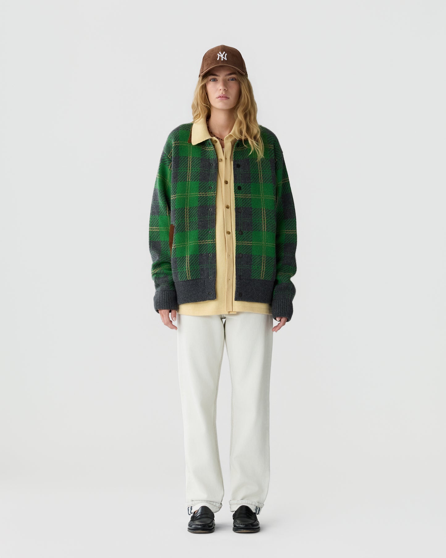 Young woman wearing a brown baseball cap, green and gray plaid cardigan over a yellow shirt, white pants, and black shoes, standing against a plain white background.