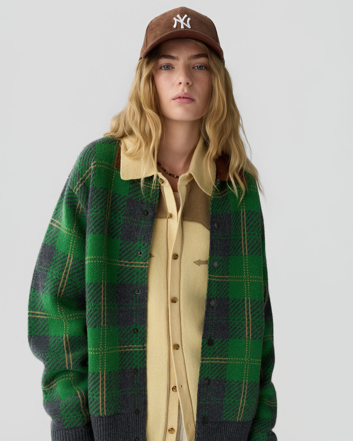 Young woman with long blonde hair wearing a brown baseball cap, beige button-up shirt, and a green and gray plaid jacket against a plain light background.