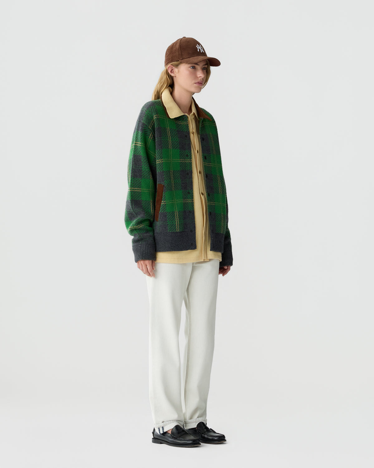 A person wearing a brown baseball cap, a green plaid cardigan over a beige shirt, white pants, and black shoes standing against a plain light gray background.