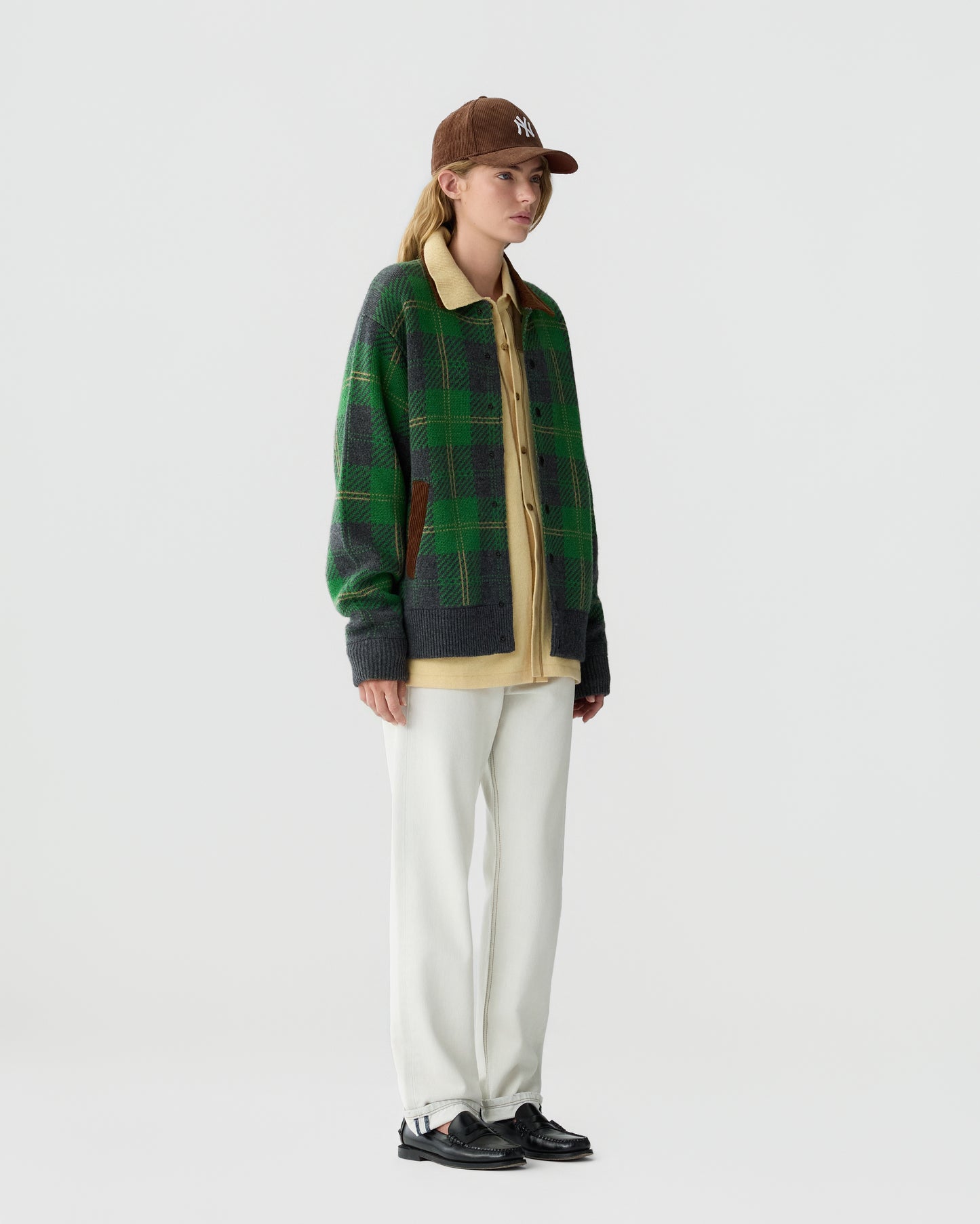 A person wearing a brown baseball cap, a green plaid cardigan over a beige shirt, white pants, and black shoes standing against a plain light gray background.