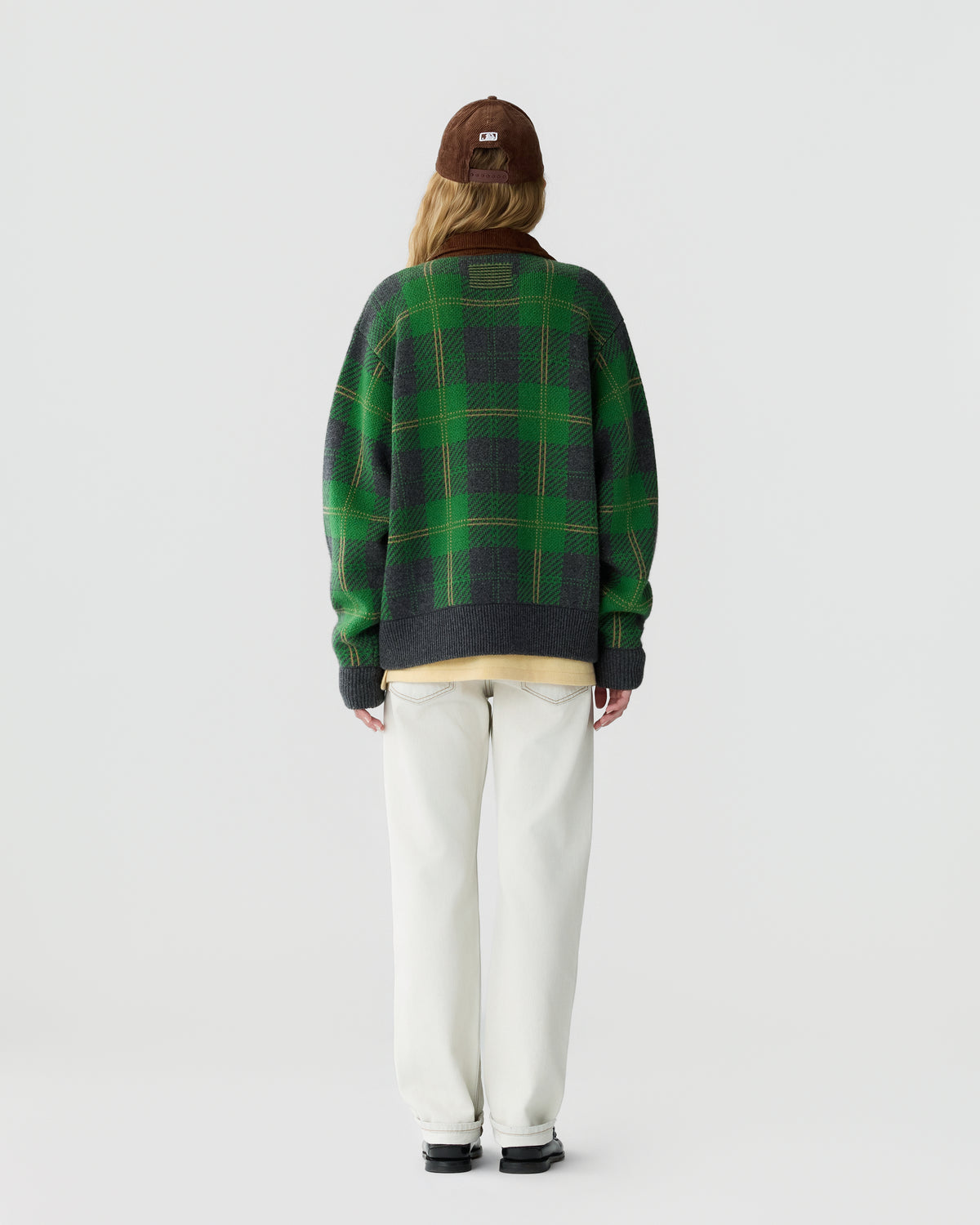 Person wearing a green and dark gray plaid sweater, off-white pants, black shoes, and a brown cap, standing facing away against a plain white background.