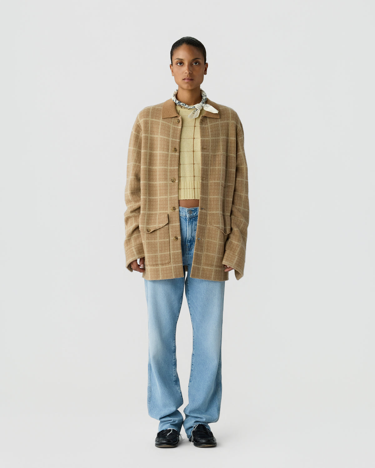 Woman wearing a beige plaid oversized button-up jacket over a yellow knit sweater, light blue jeans, black shoes, and a patterned neck scarf, standing against a plain background.