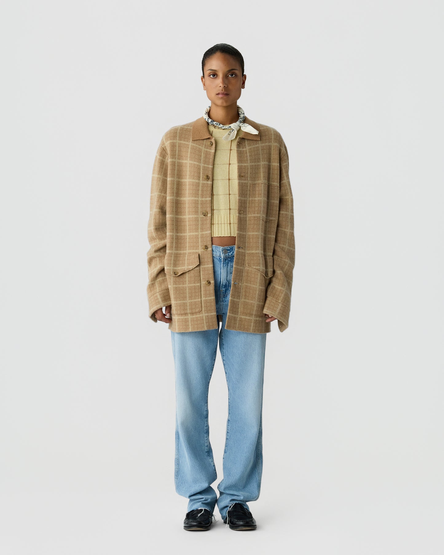 Woman wearing a beige plaid oversized button-up jacket over a yellow knit sweater, light blue jeans, black shoes, and a patterned neck scarf, standing against a plain background.