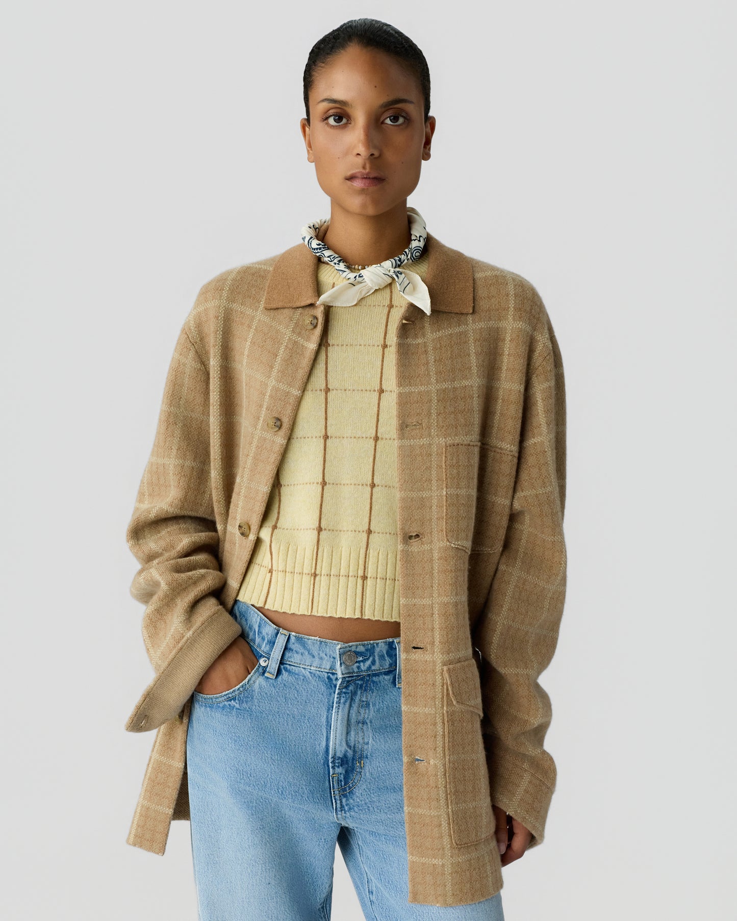Woman wearing a beige plaid jacket over a light yellow cropped sweater with a checkered pattern, paired with light blue jeans and a white scarf tied around her neck.