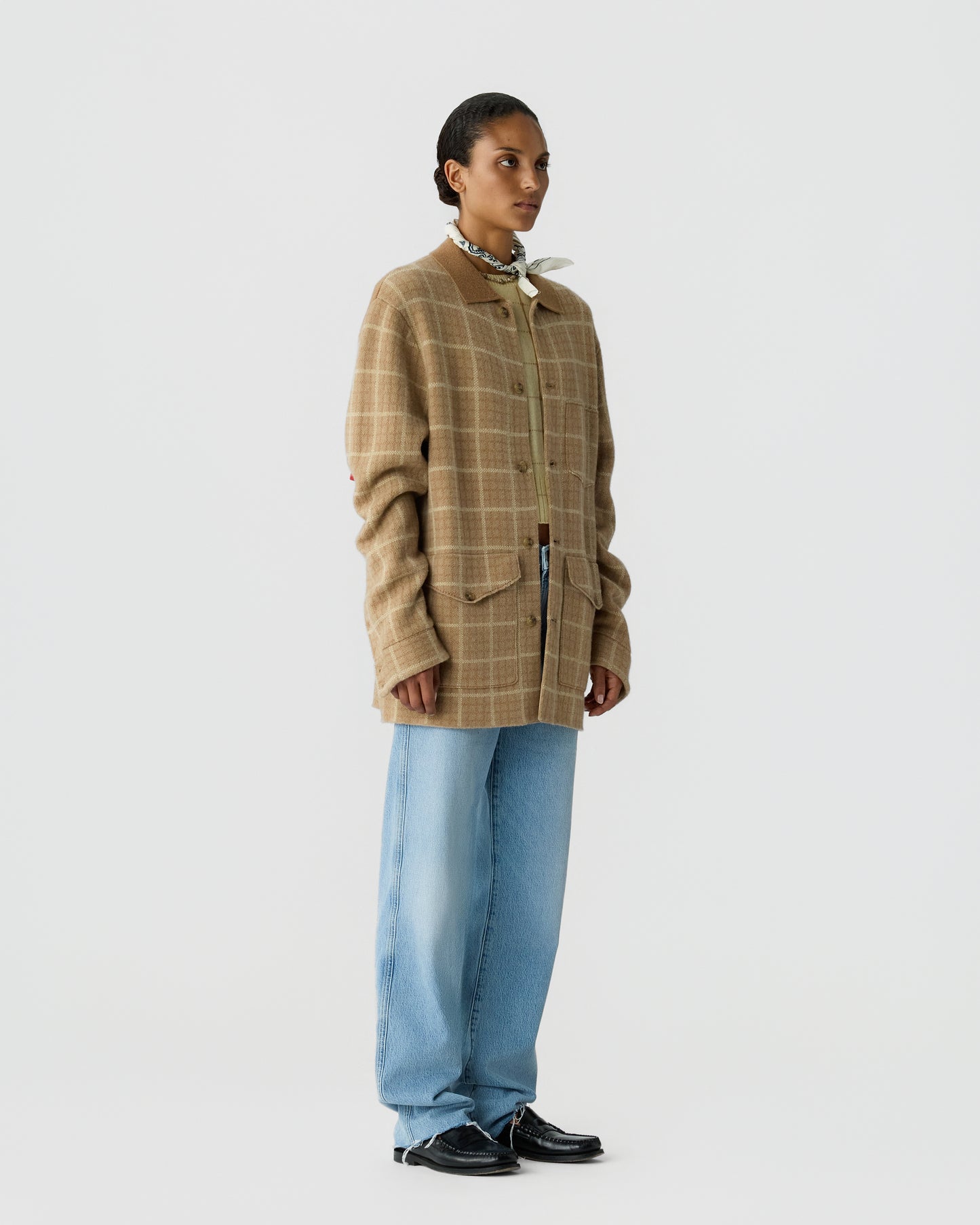 Woman wearing a beige plaid oversized jacket with a brown collar, light blue wide-leg jeans, black shoes, and a small scarf tied around her neck against a plain white background.
