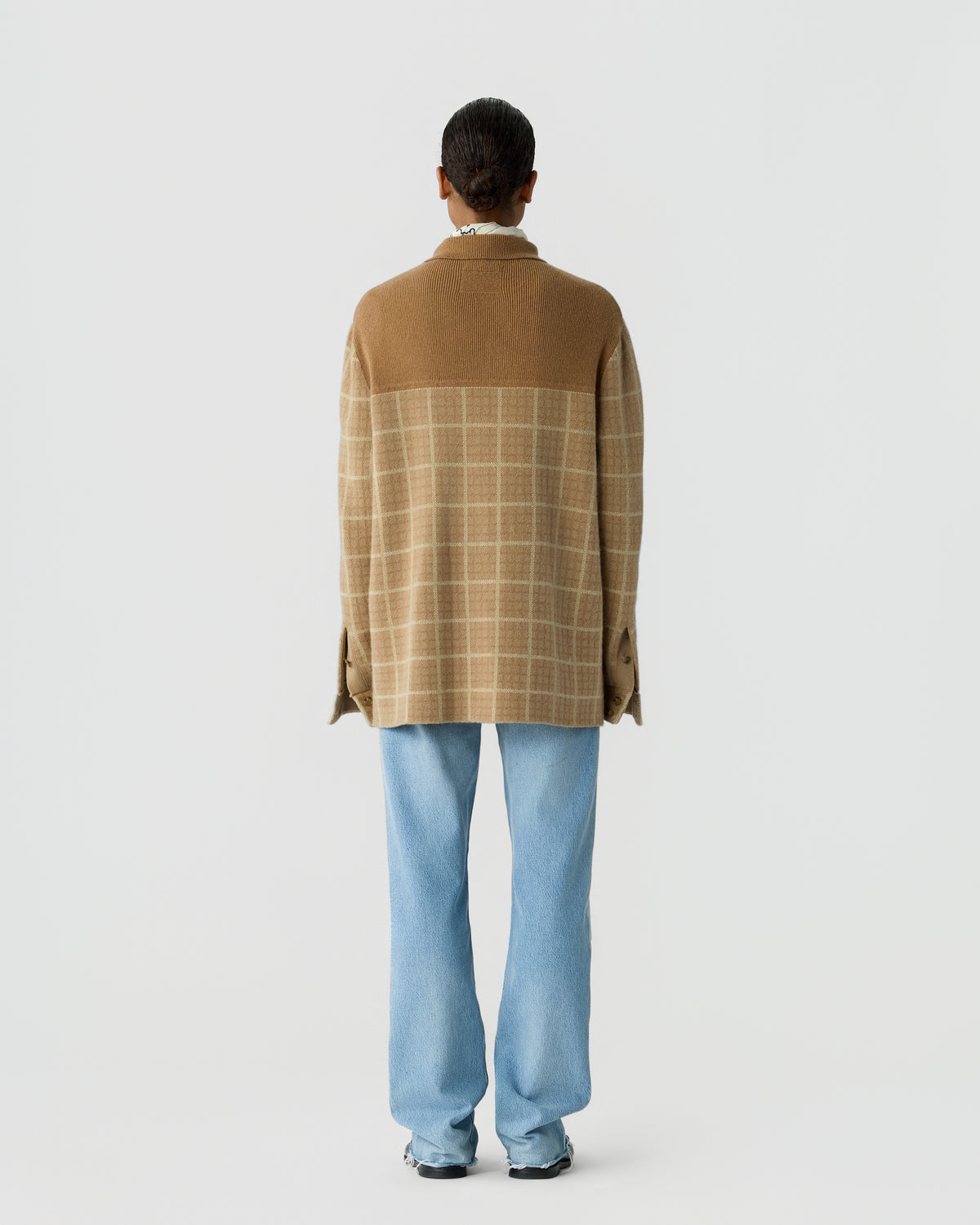 Person standing with their back to the camera wearing a brown and beige checked cashmere sweater and light blue jeans against a plain white background.